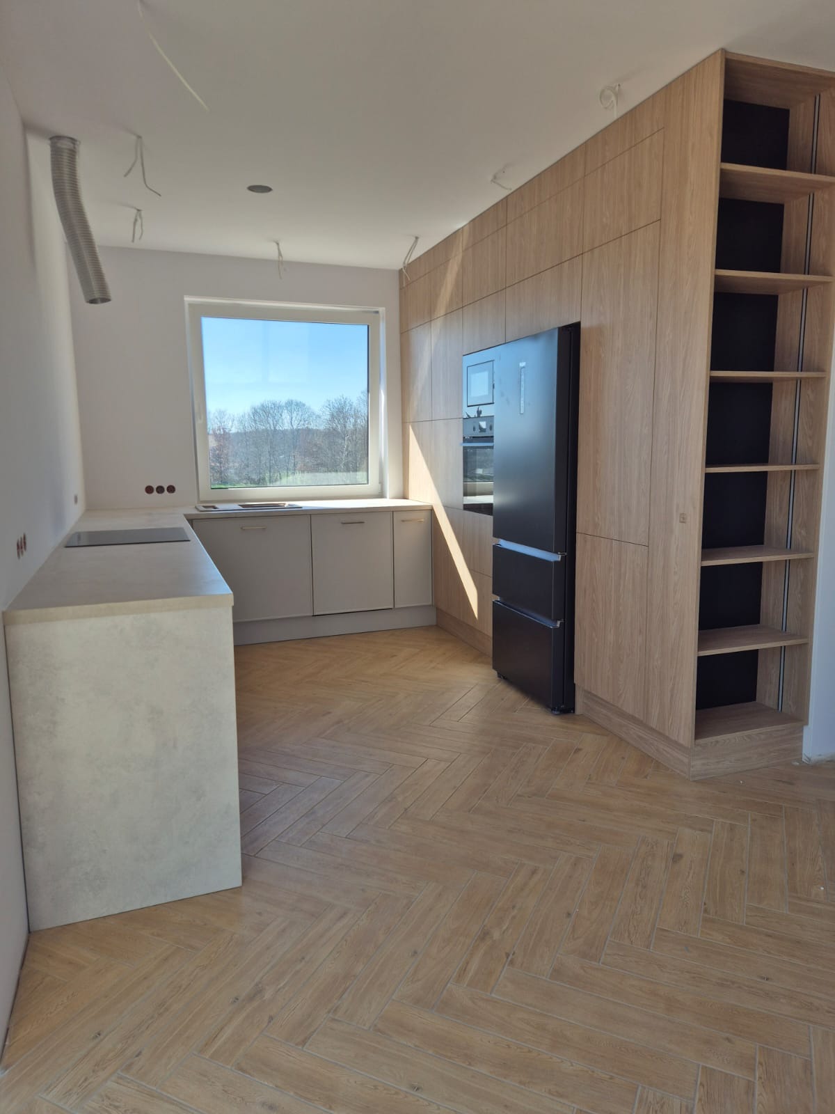 Jasna kuchnia z drewnianymi frontami, czarną lodówką i parkietem ułożonym w jodełkę. Widoczne niedokończone elementy instalacji na suficie.
