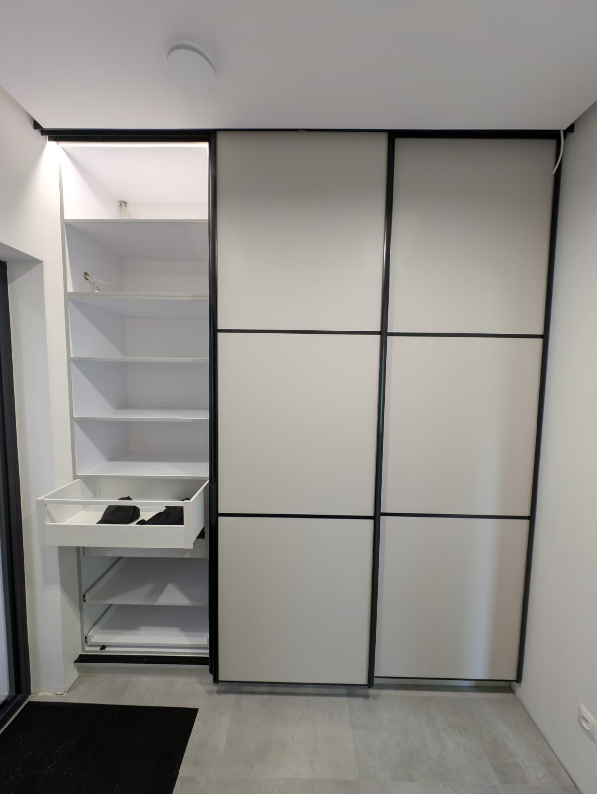 Biała szafa wnękowa z przesuwnymi drzwiami i czarnymi ramami. Jedna część szafy otwarta, widoczne półki i wysuwana szuflada z ubraniami. Szara podłoga.