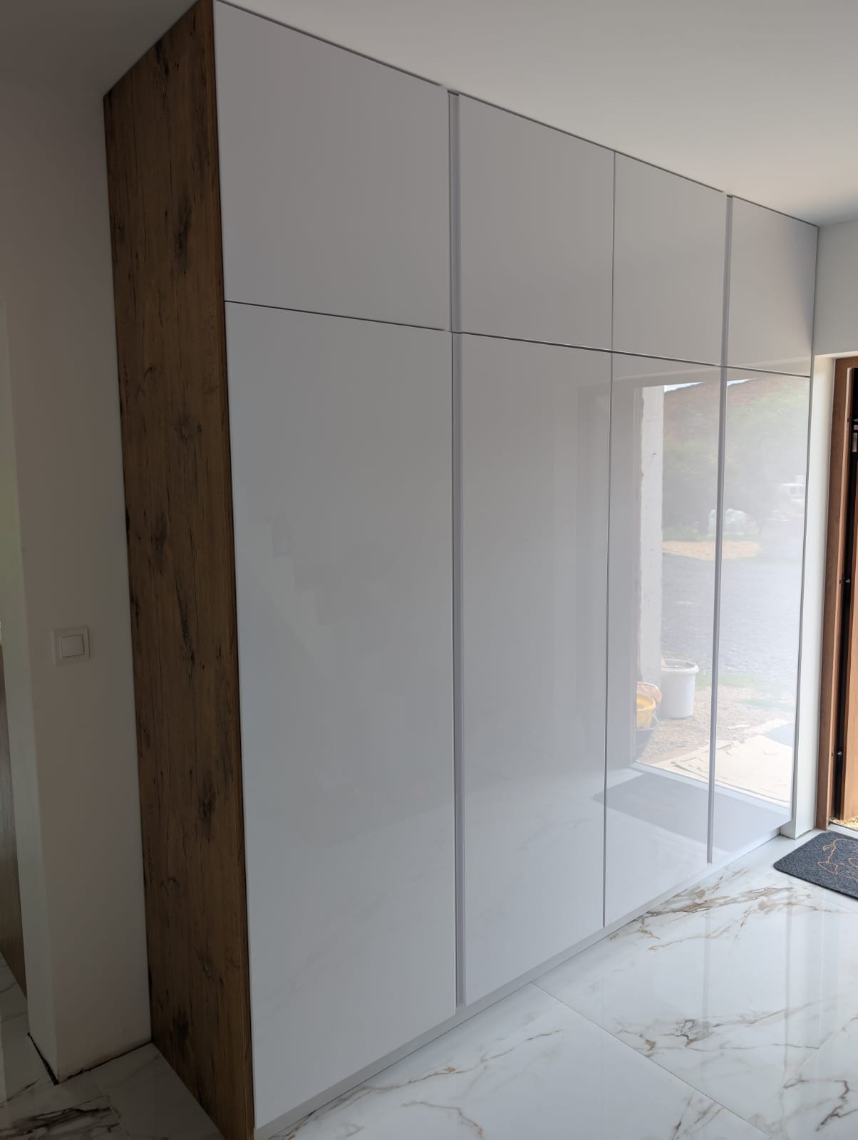Biała szafa wnękowa z drewnianym panelem bocznym, błyszczące fronty, marmurowa podłoga, widok na zewnątrz przez szparę między drzwiami a szafą.