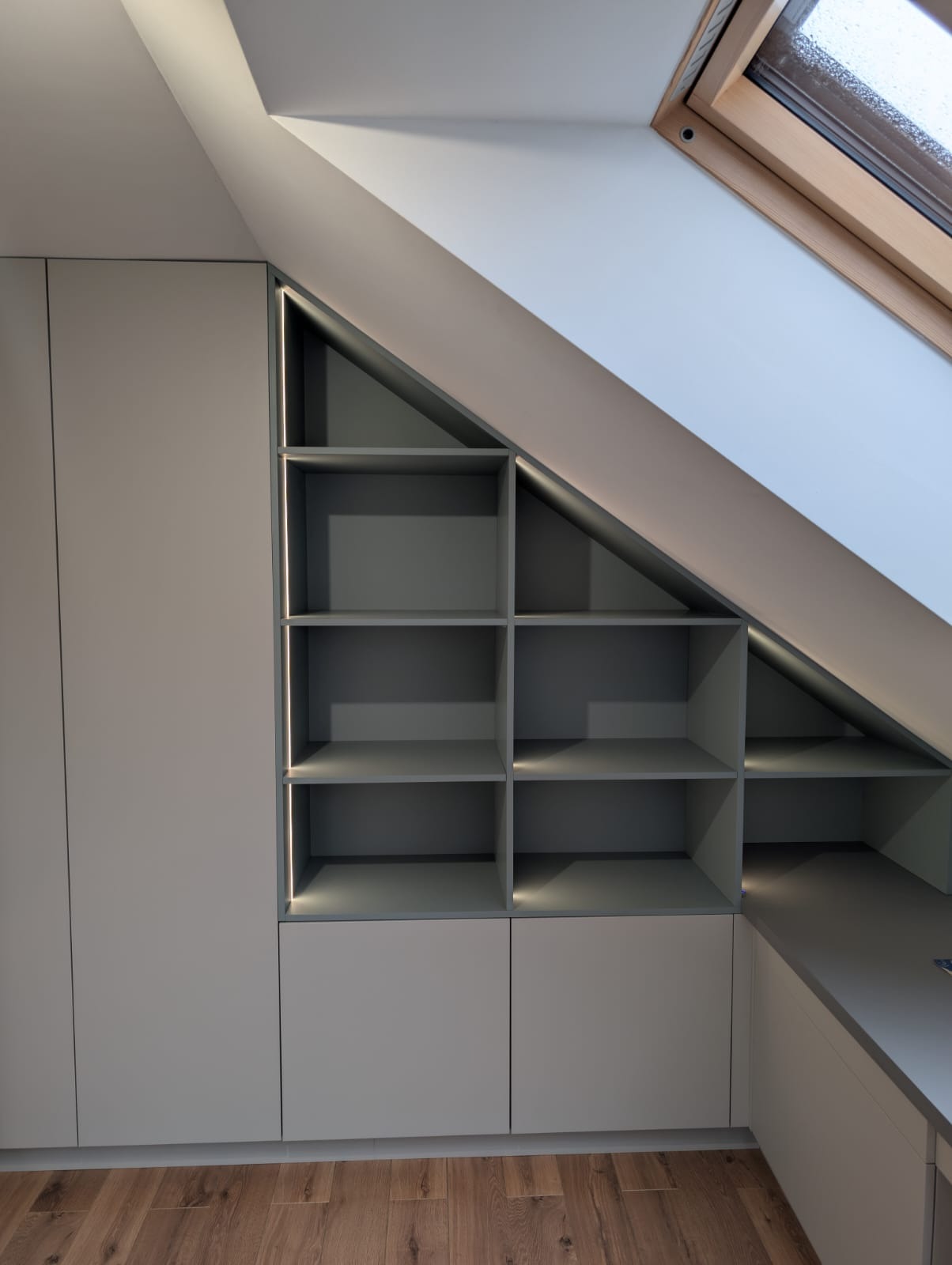 Szara szafa wnękowa z półkami pod skosem, oświetlenie LED, drewniana podłoga i okno dachowe. Minimalistyczny design.