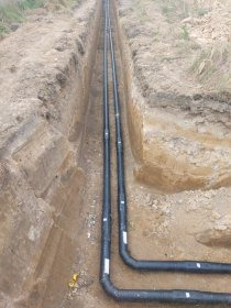 Dwie równoległe rury wodno-kanalizacyjne w wykopie ziemnym, z widocznymi złączkami i oznaczeniami, gotowe do zasypania. Jasna ziemia, naturalne światło.