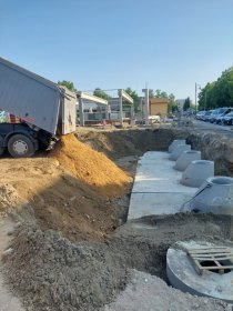 Wykop z ułożonymi betonowymi studniami kanalizacyjnymi, zasypywany piaskiem z ciężarówki. W tle budowa i drzewa.