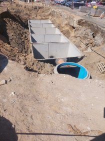 Betonowe studnie kanalizacyjne w wykopie, widoczne rury przyłączeniowe. Etap budowy sieci wodno-kanalizacyjnej, paleta w tle.