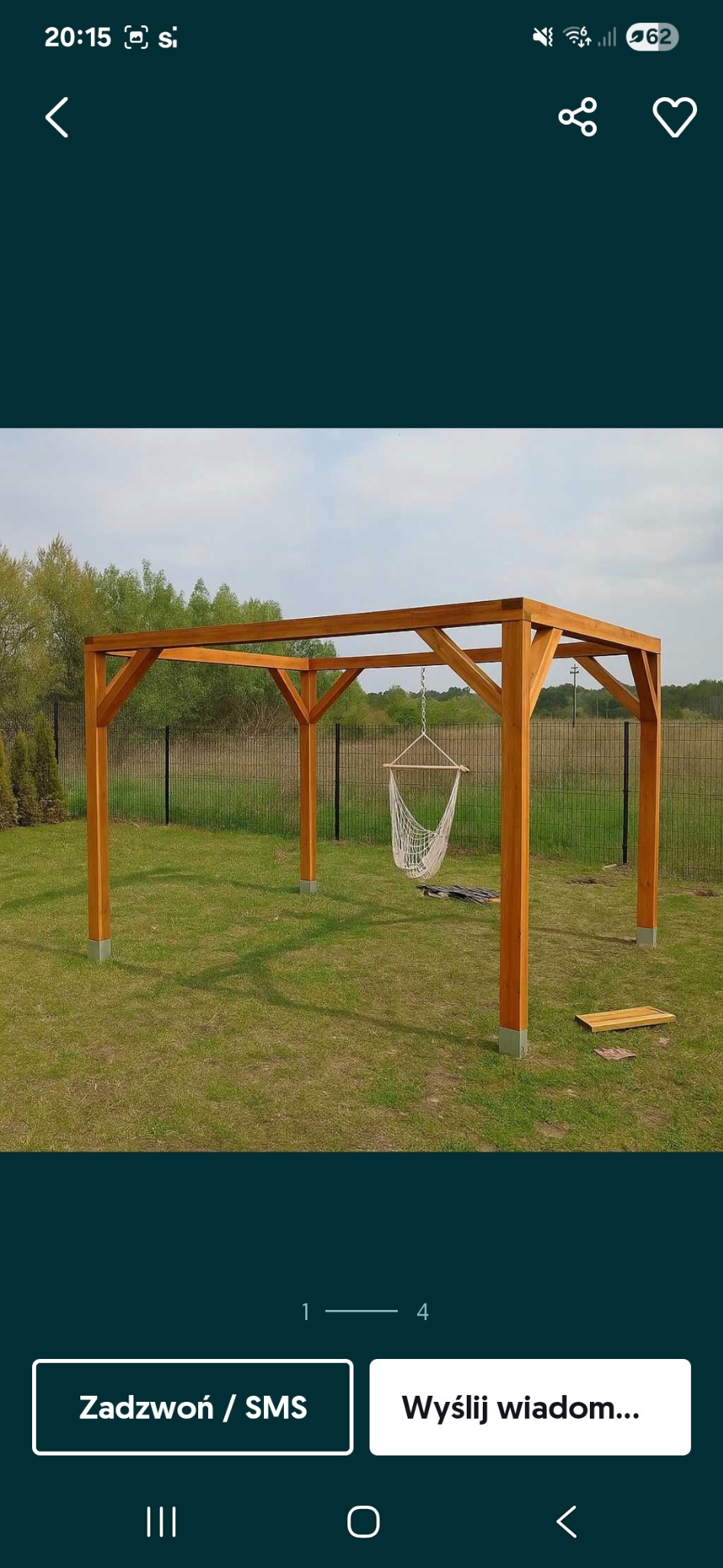 Drewniana pergola z wiszącym hamakiem na tle zielonego trawnika i ogrodzenia z siatki. Elementy montażowe widoczne na ziemi.