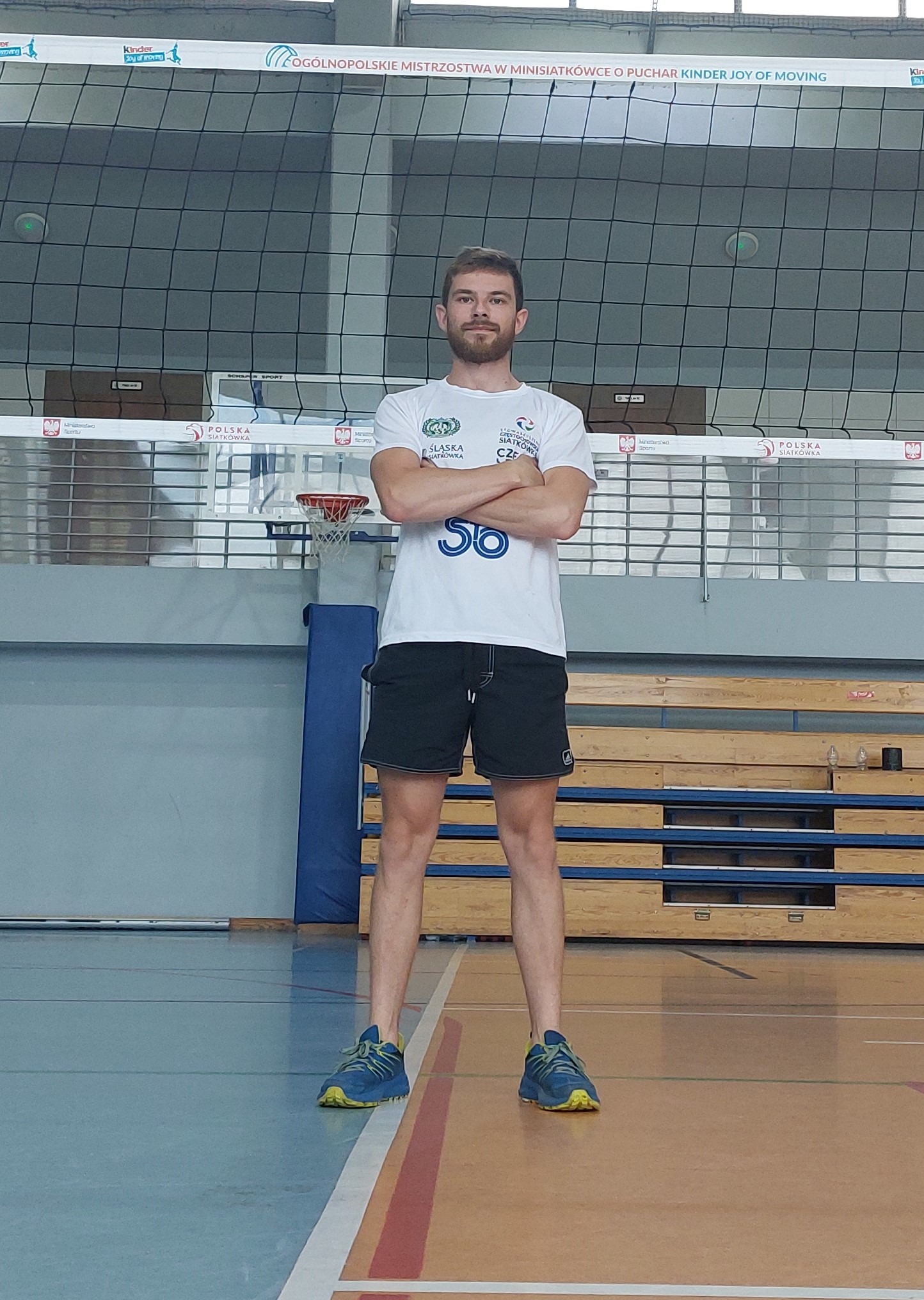 Młody mężczyzna w sportowym stroju stoi na tle siatki do siatkówki, z założonymi rękami. Hala sportowa z koszem do koszykówki w tle.
