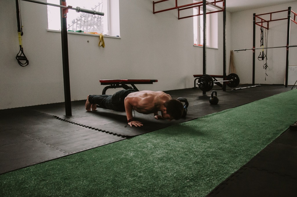 Mężczyzna w trakcie pompek na podłodze w siłowni, widoczny sprzęt do ćwiczeń i sztuczna trawa. Osoba bez koszulki, widoczne mięśnie. Trening w jasnym pomieszczeniu.
