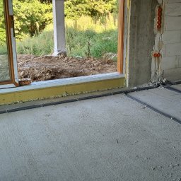 MR-Elektro - Instalacja elektryczna podtynkowa w nowym budynku, widoczne rury karbowane w podłodze i puszki w ścianie z betonu komórkowego. W tle okno z widokiem na zewnątrz.