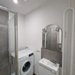 Nowoczesna łazienka z kabiną prysznicową, pralką Finlux i umywalką z szafką. Szaro-biała kolorystyka, oświetlenie punktowe i owalne lustro z kinkietami.