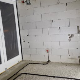 MR-Elektro - Surowa ściana z betonu komórkowego z widoczną instalacją elektryczną podtynkową, obok okno z folią ochronną. W tle widoczny element oświetlenia.