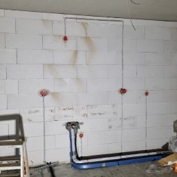 MR-Elektro - Surowy mur z instalacją elektryczną i rurami w nieotynkowanym pomieszczeniu. Widoczne kable i puszki elektryczne, rury w niebieskiej izolacji. W tle drabina i sprzęt budowlany.
