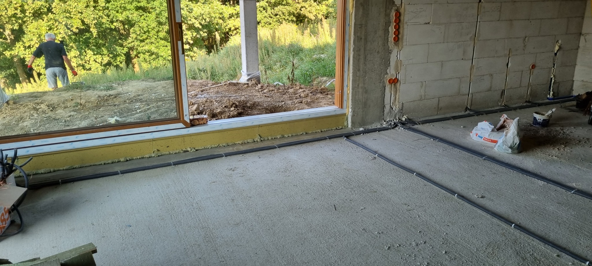 Instalacja elektryczna podtynkowa w nowym budynku, widoczne rury karbowane w podłodze i puszki w ścianie z betonu komórkowego. W tle okno z widokiem na zewnątrz.
