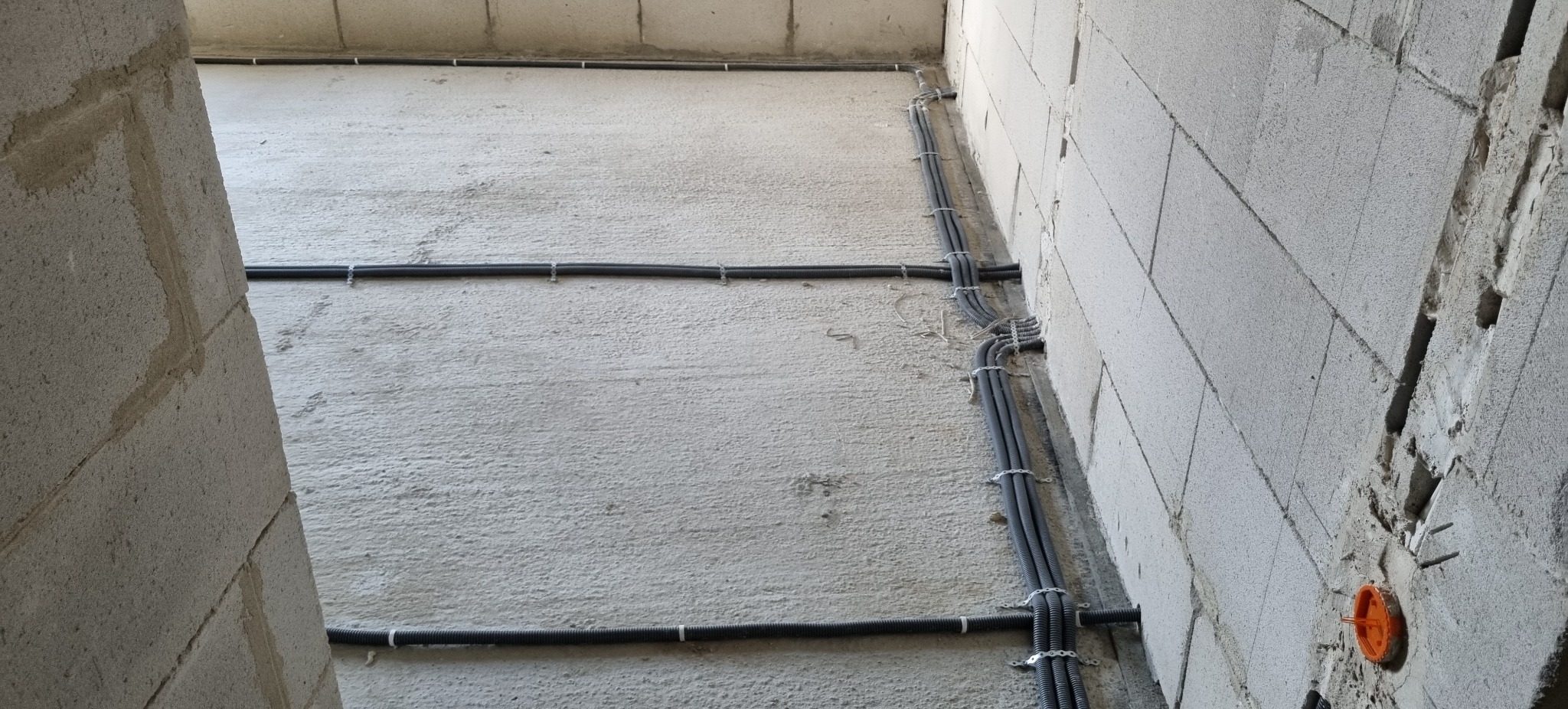 Szare rury instalacji elektrycznej, przymocowane klamrami do surowej ściany z betonu komórkowego. Widoczne gniazdo elektryczne w ścianie.