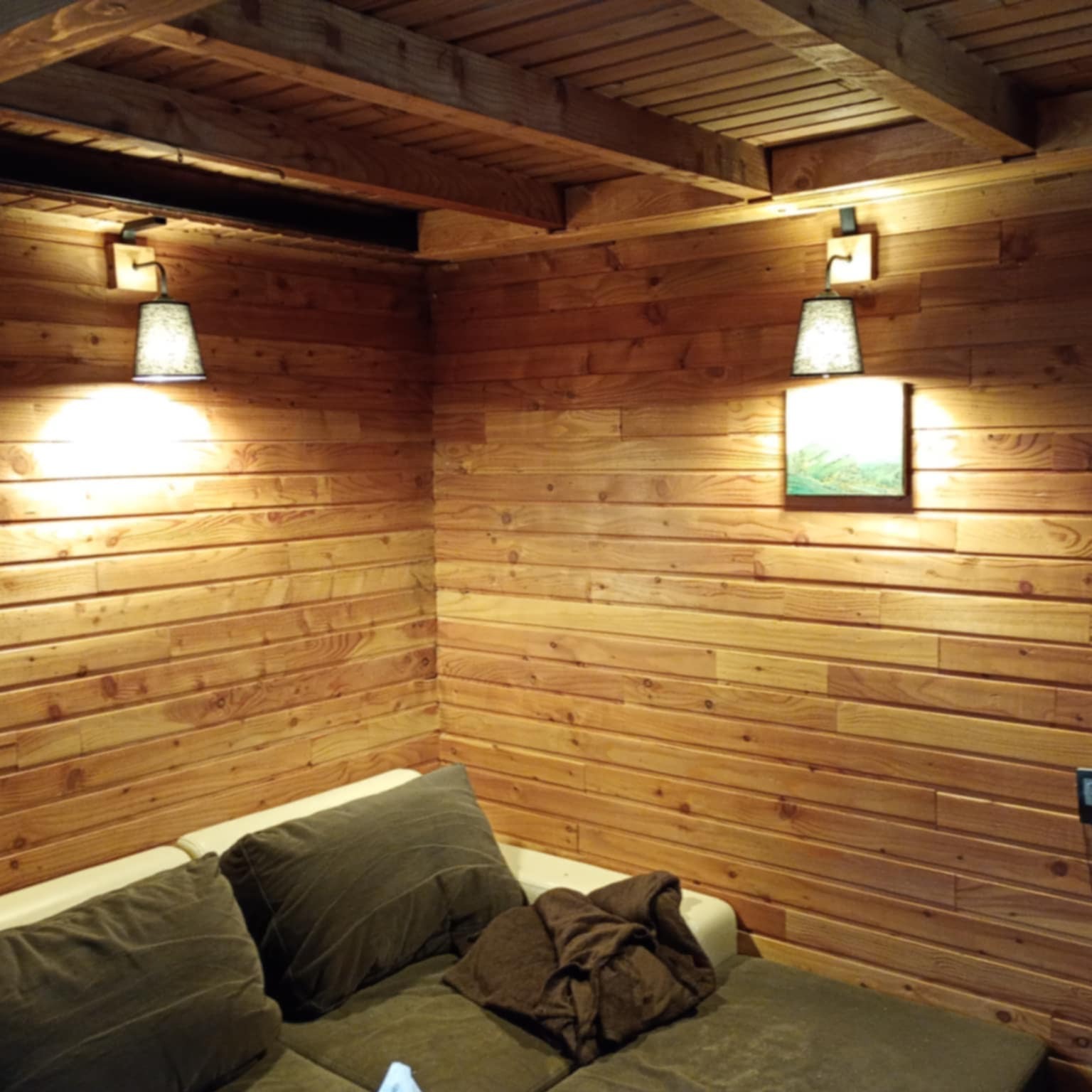 Drewniane wnętrze z kanapą, oświetlone dwoma lampami ściennymi. Widoczne drewniane ściany i sufit, tworzące przytulny klimat. Dekoracyjne oświetlenie.
