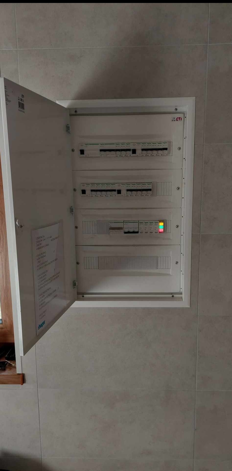 Otwarta skrzynka elektryczna ETI w ścianie z szarych płytek, widoczne bezpieczniki i dokumentacja wewnątrz. Dolny rząd z diodami sygnalizacyjnymi.