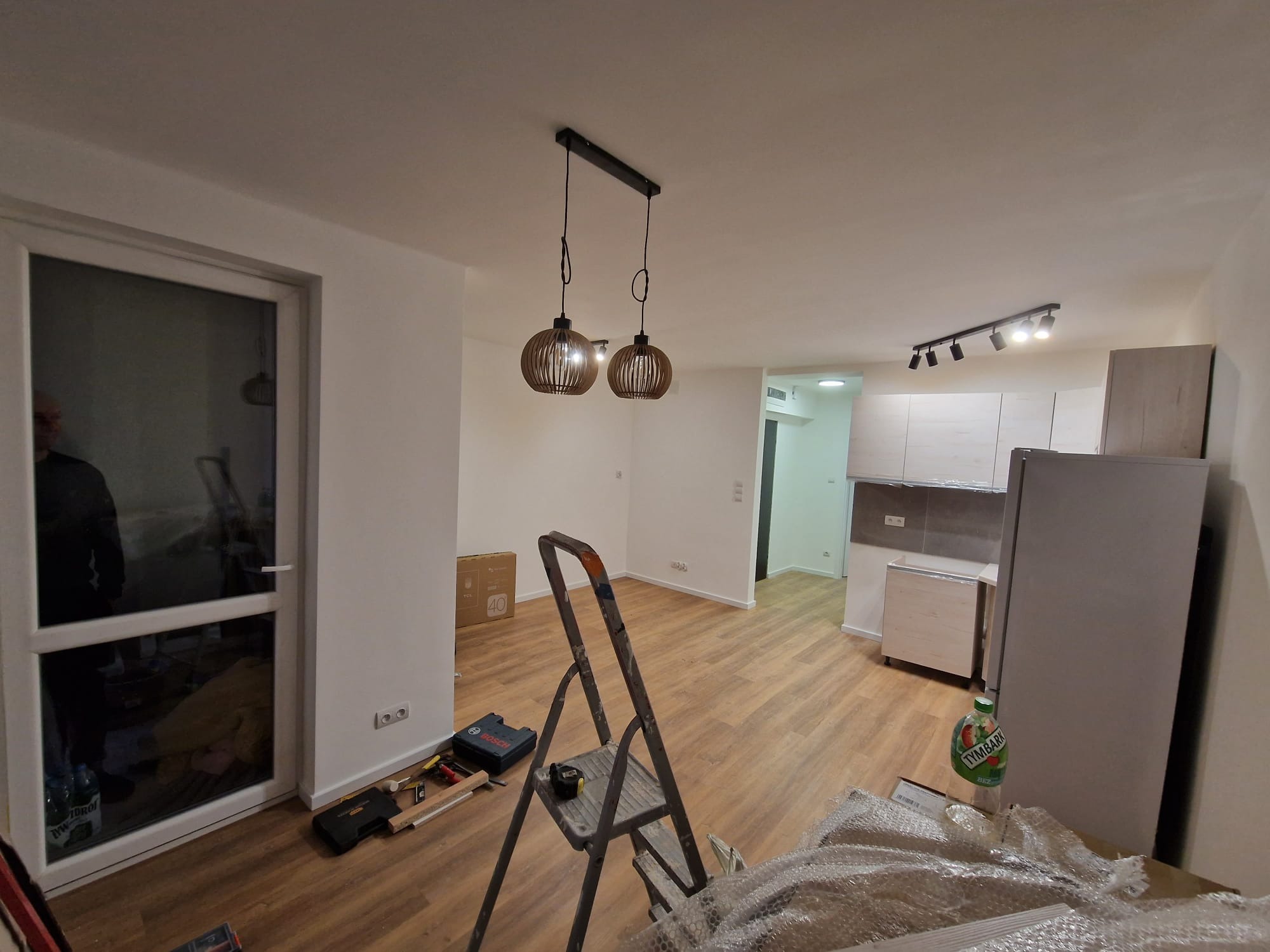 Wnętrze mieszkania w trakcie remontu: drabina, narzędzia, częściowo rozpakowane meble kuchenne, nowe oświetlenie sufitowe. Widok na odnowioną przestrzeń.