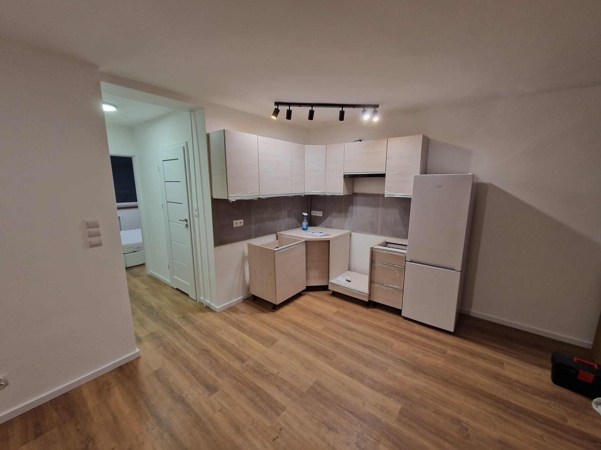 Jasna kuchnia w trakcie montażu: widoczne szafki, blat narożny, lodówka i listwa oświetleniowa. Podłoga drewniana, w tle otwarte drzwi do innego pomieszczenia.