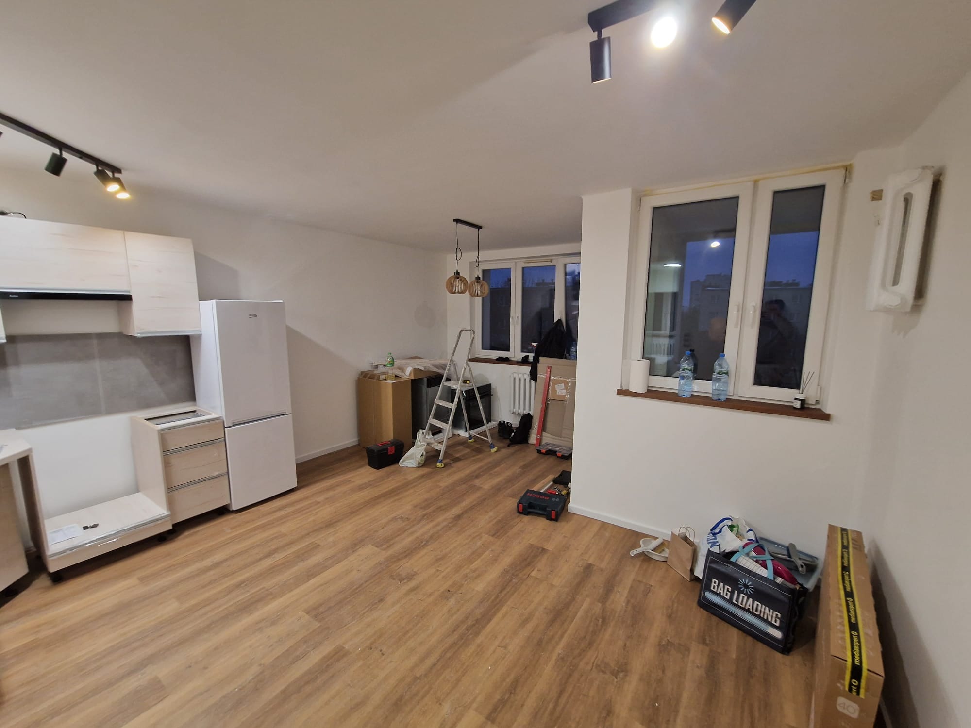 Wnętrze mieszkania w trakcie remontu: częściowo zamontowana kuchnia, podłoga, drabina, narzędzia. Widoczne elementy instalacji elektrycznej.