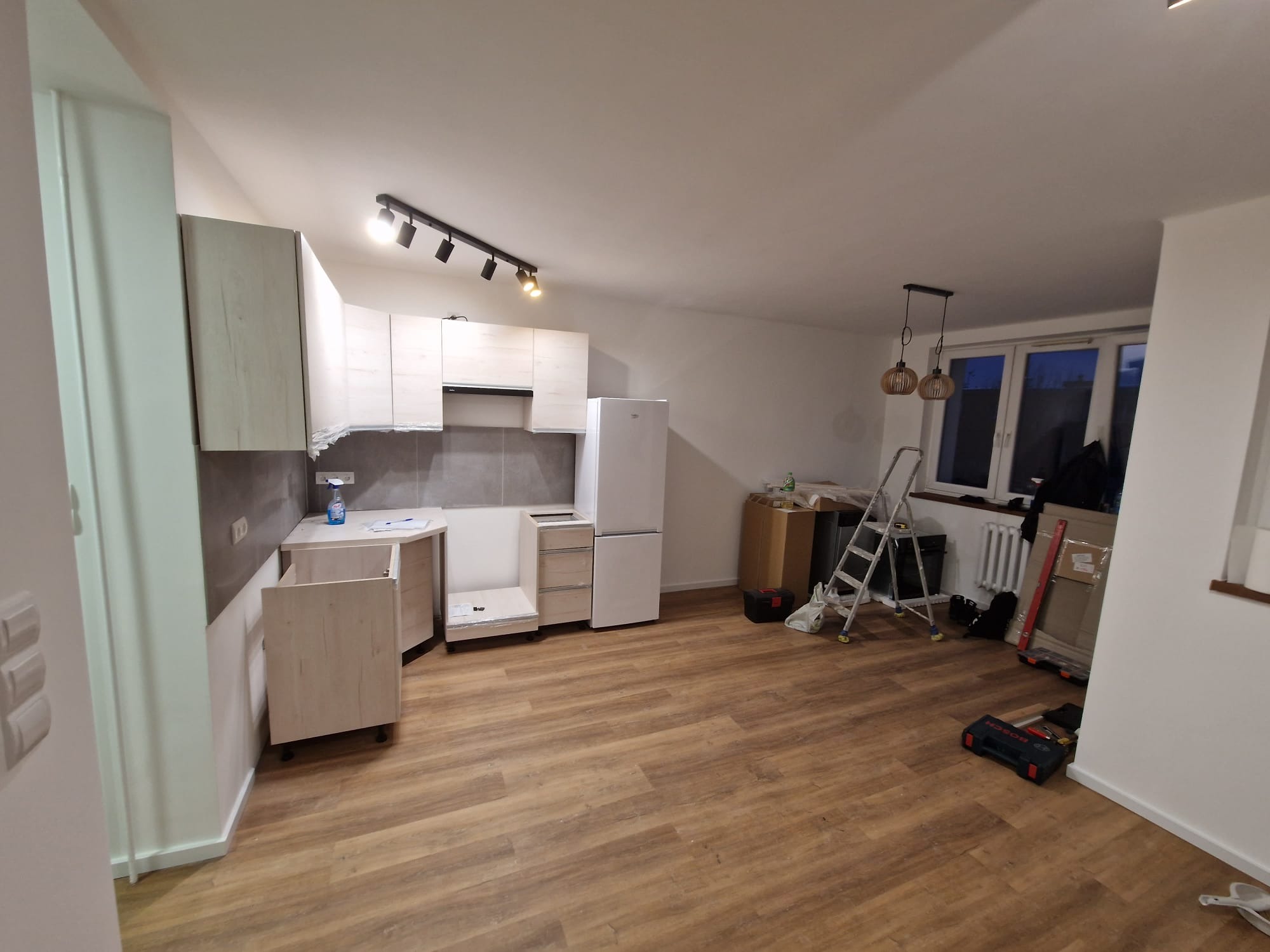 Częściowo zmontowana, jasna kuchnia z drewnopodobną podłogą, widok na okno i narzędzia, sugerujący etap prac remontowych. Oświetlenie sufitowe punktowe.