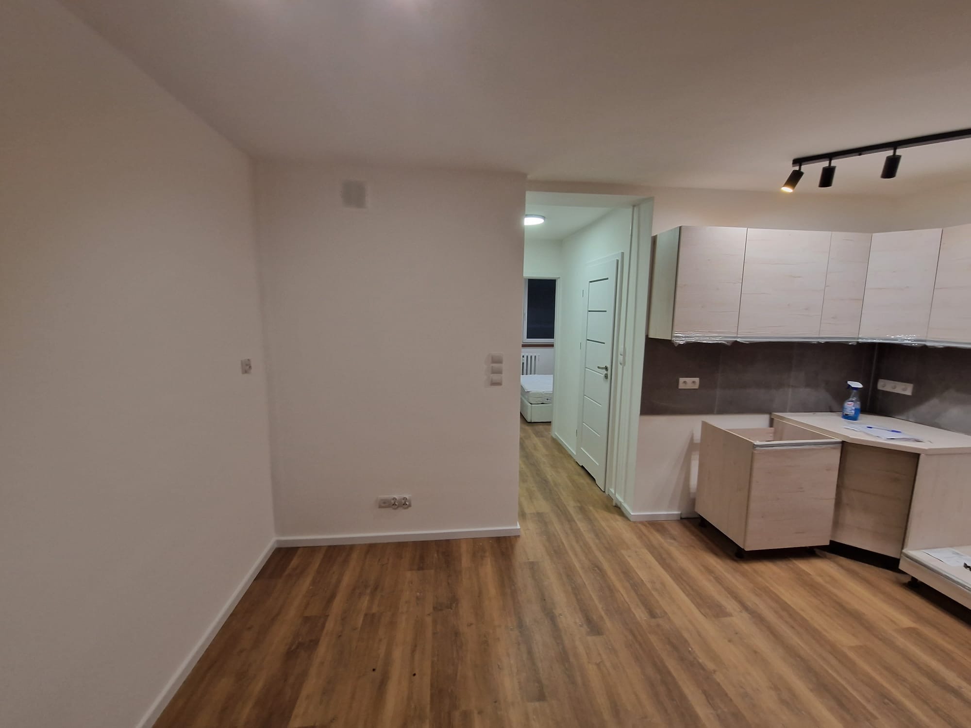 Wnętrze mieszkania w trakcie remontu: częściowo zmontowana jasna kuchnia, surowe ściany, parkiet, widok na pokój. Oświetlenie szynowe.