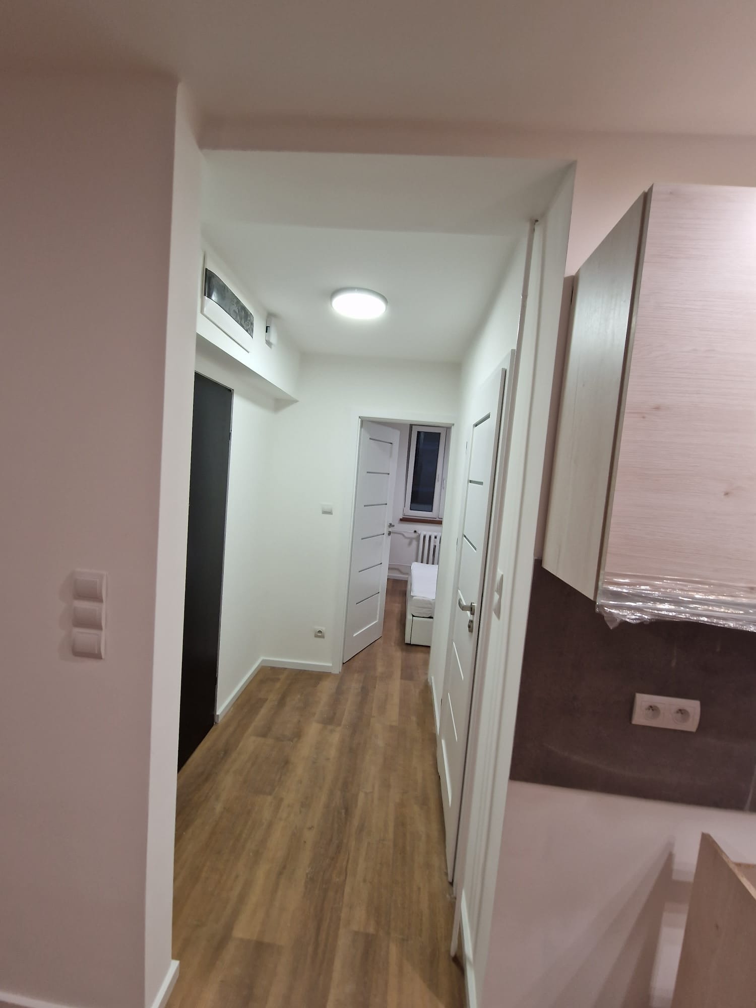 Nowoczesne wnętrze: jasny korytarz z drewnianą podłogą, białymi ścianami i drzwiami. Widoczne gniazdka elektryczne i fragment mebli kuchennych.