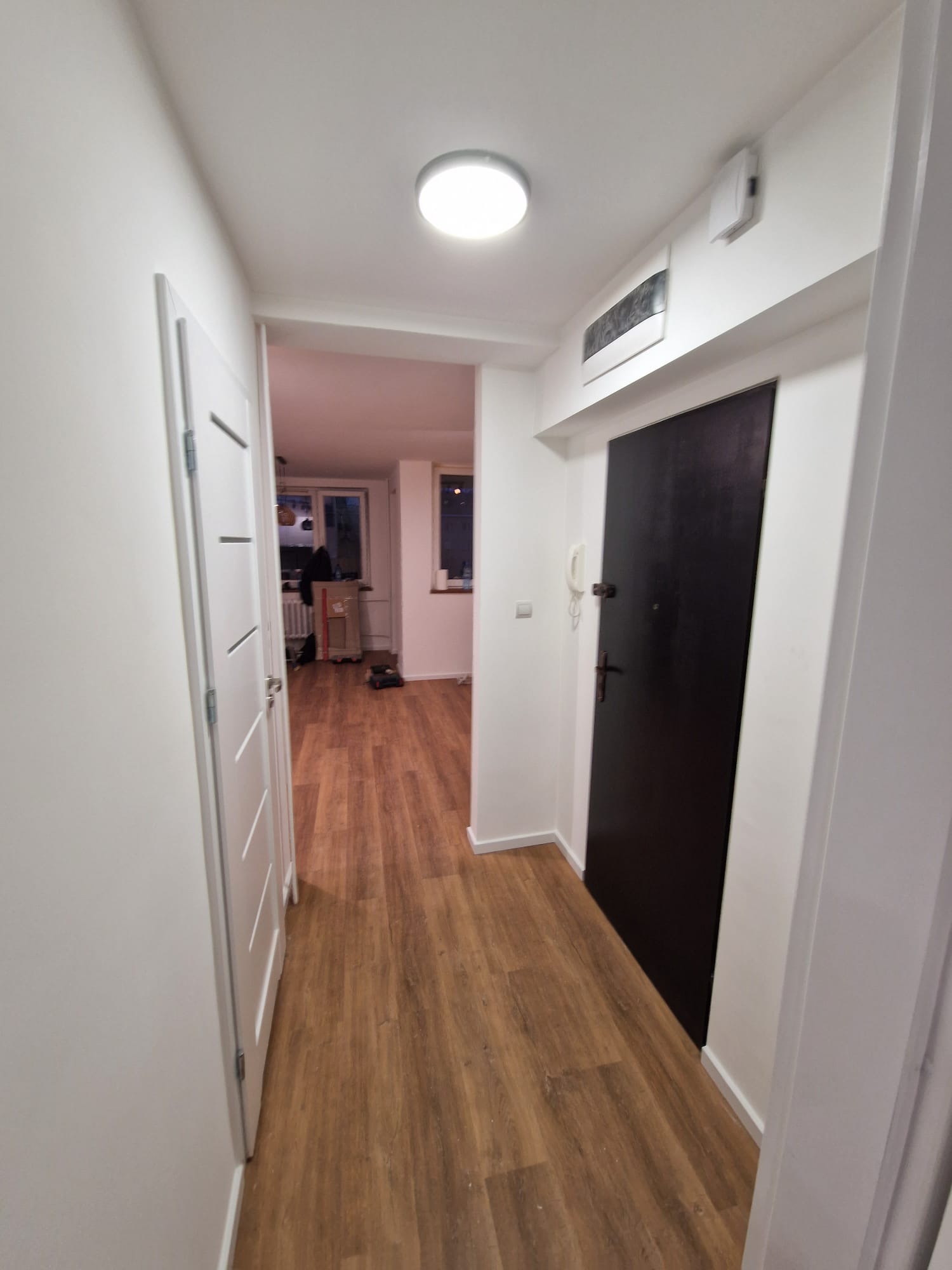 Nowoczesny korytarz po remoncie: panele podłogowe imitujące drewno, białe ściany i drzwi, czarne drzwi wejściowe z domofonem. Widok na dalszą część mieszkania.