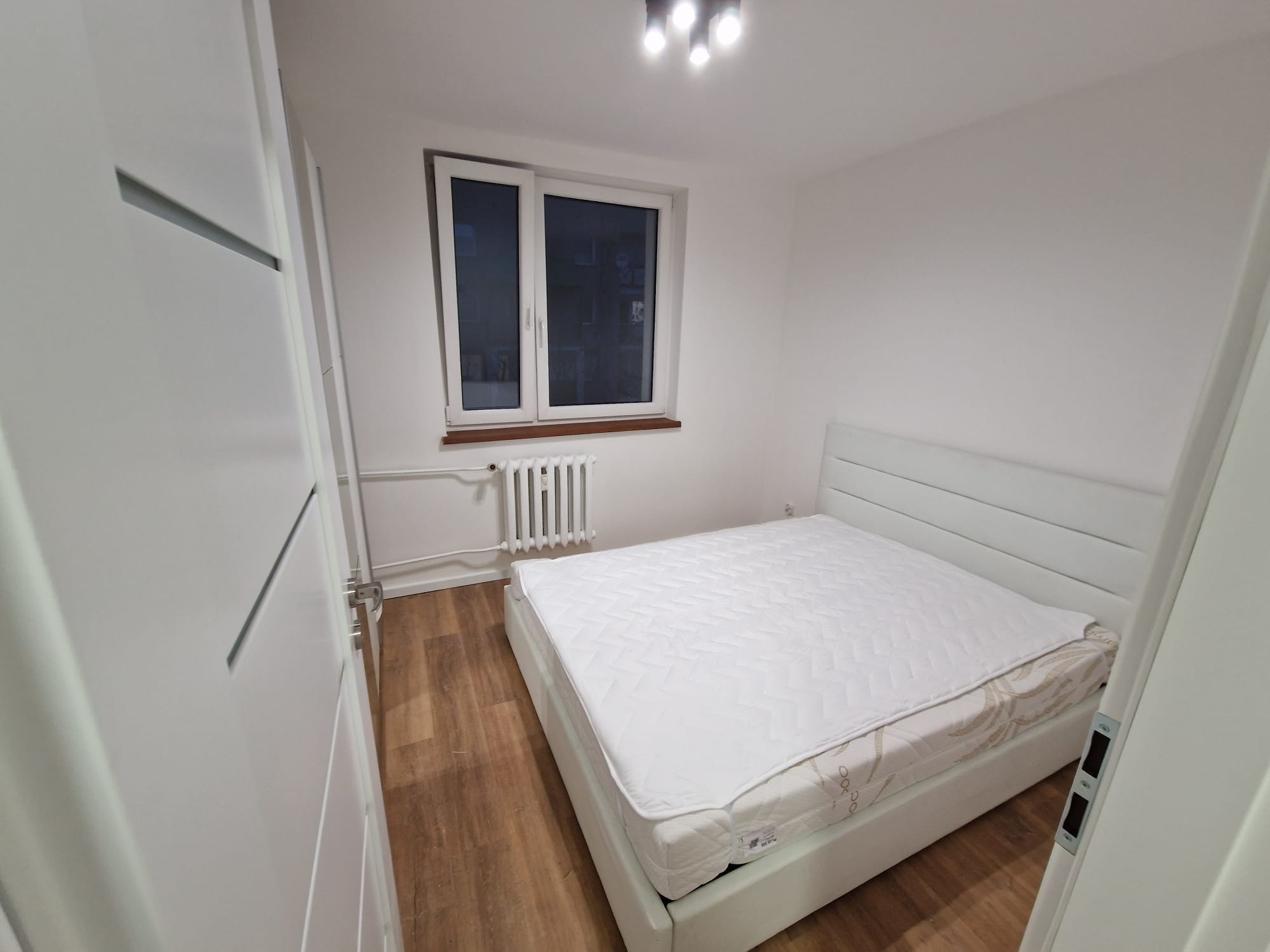 Jasne wnętrze sypialni po remoncie: białe ściany, łóżko z materacem i grzejnik pod oknem. Widok zza uchylonych drzwi.