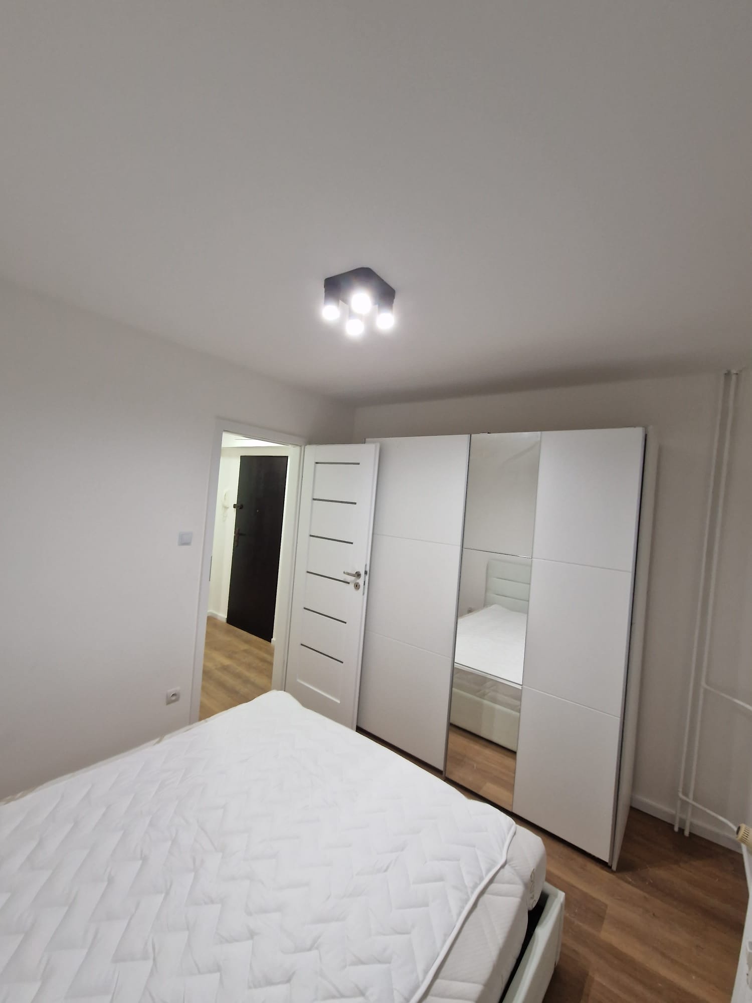 Jasne wnętrze sypialni po remoncie: białe ściany, drewniana podłoga, łóżko z białą pościelą i duża szafa z lustrami. Widok na drzwi.