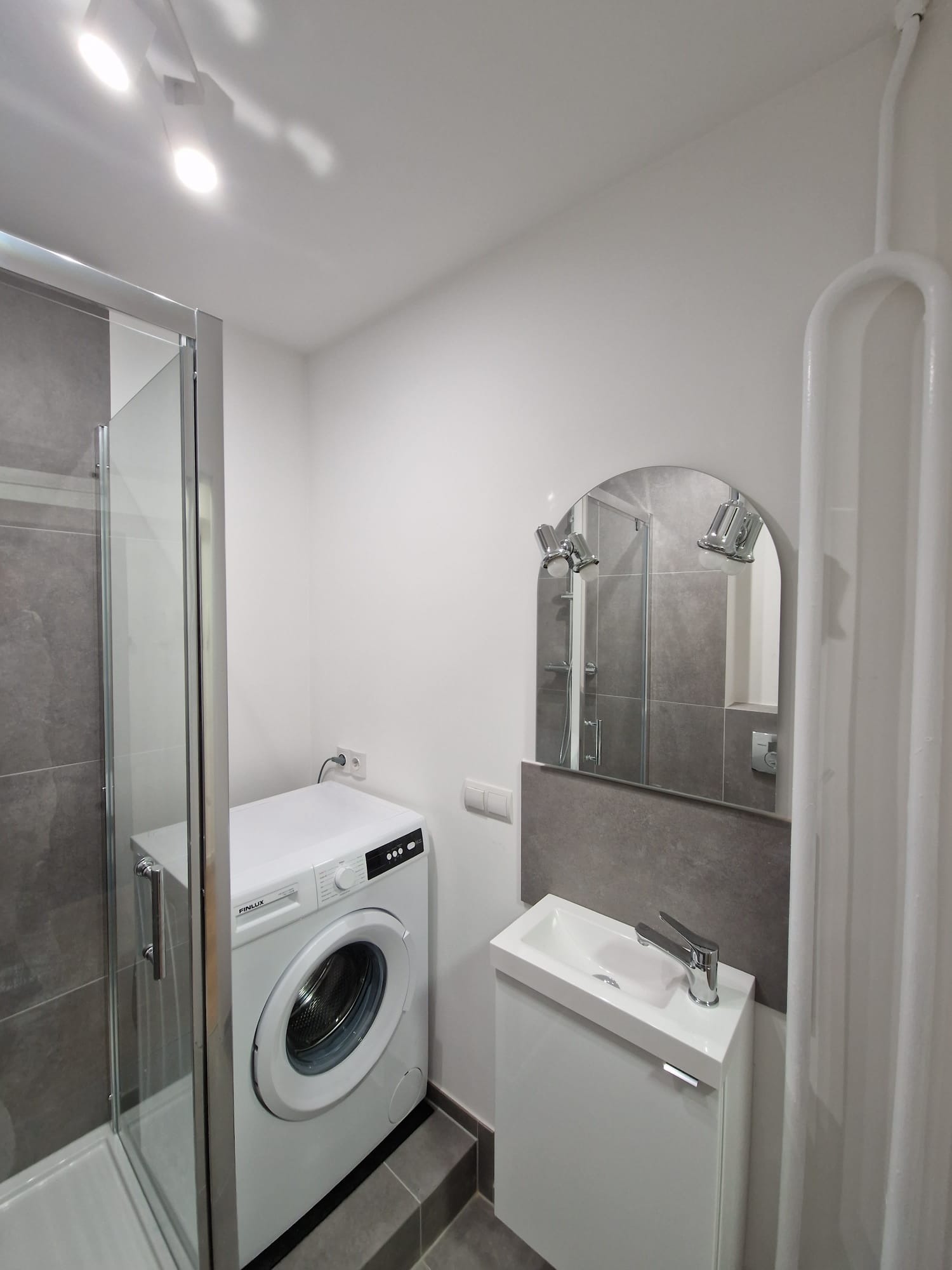 Nowoczesna łazienka z kabiną prysznicową, pralką Finlux i umywalką z szafką. Szaro-biała kolorystyka, oświetlenie punktowe i owalne lustro z kinkietami.