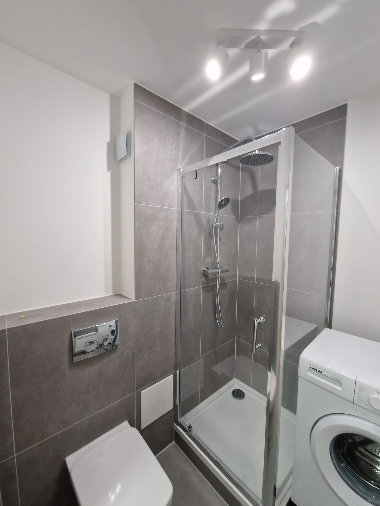 Nowoczesna łazienka po remoncie: szare płytki, kabina prysznicowa ze srebrnymi profilami, biała toaleta i pralka. Oświetlenie punktowe.