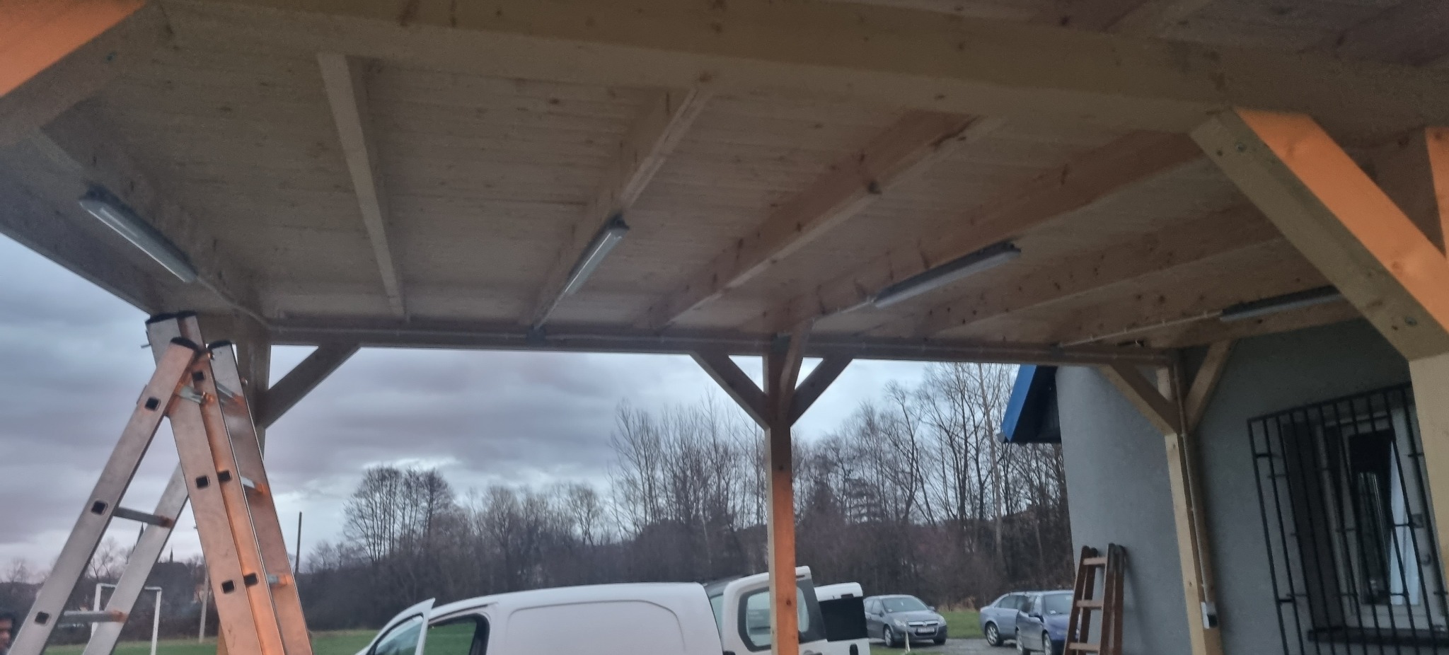 Drewniana konstrukcja z zamontowanym oświetleniem LED, widoczna drabina i samochody w tle. Oświetlenie pod zadaszeniem.