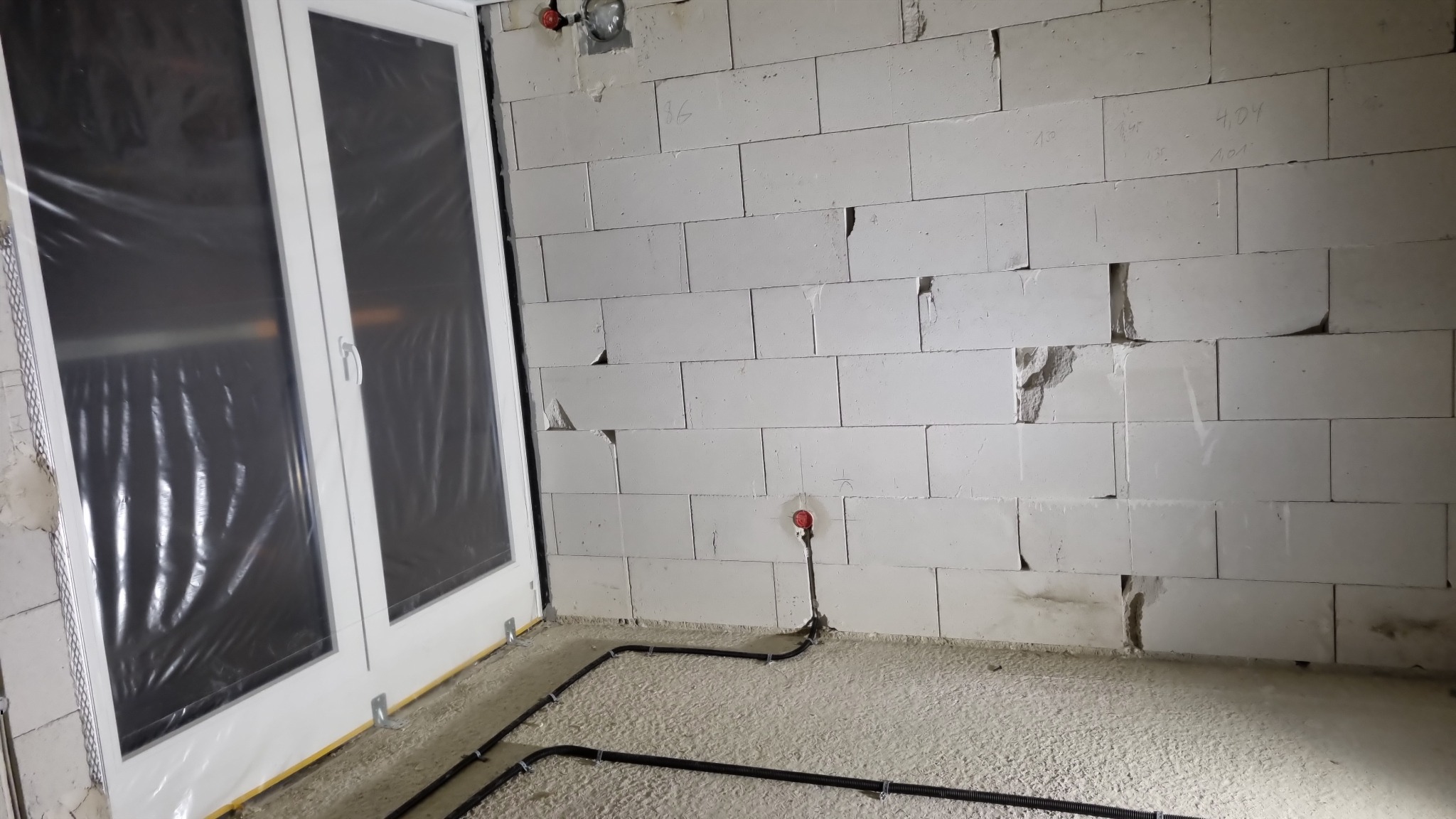 Surowa ściana z betonu komórkowego z widoczną instalacją elektryczną podtynkową, obok okno z folią ochronną. W tle widoczny element oświetlenia.
