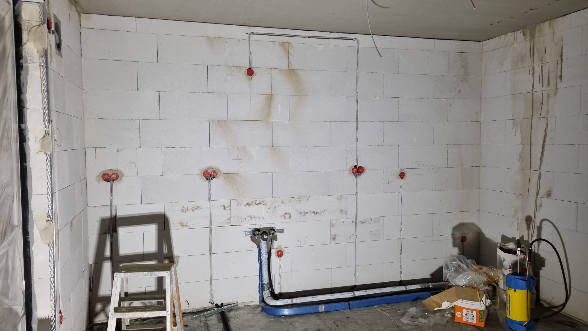 Surowy mur z instalacją elektryczną i rurami w nieotynkowanym pomieszczeniu. Widoczne kable i puszki elektryczne, rury w niebieskiej izolacji. W tle drabina i sprzęt budowlany.