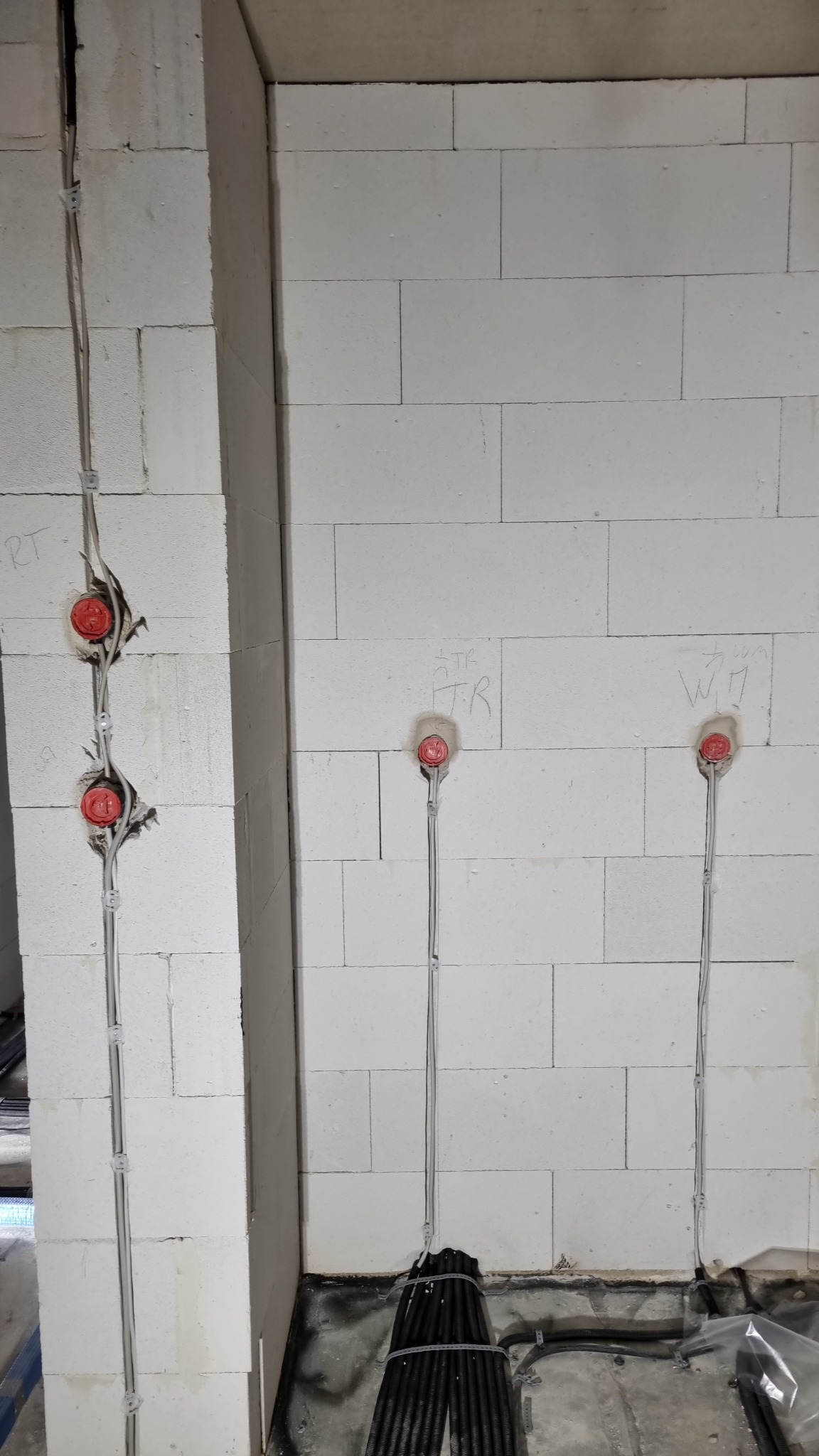 Ściana z betonu komórkowego w trakcie montażu instalacji elektrycznej, widoczne kable w peszlach i puszki podtynkowe z czerwonymi dekielkami.  Na ścianie napisy ołówkiem.