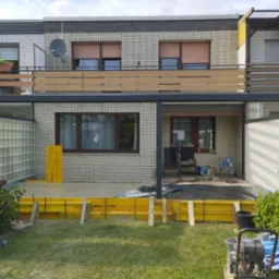 Budowa tarasu przy domu z cegły: żółte szalunki, w tle balkon z drewnianą balustradą, widoczny sprzęt budowlany i pojemnik na śmieci. Etap wylewania betonu.