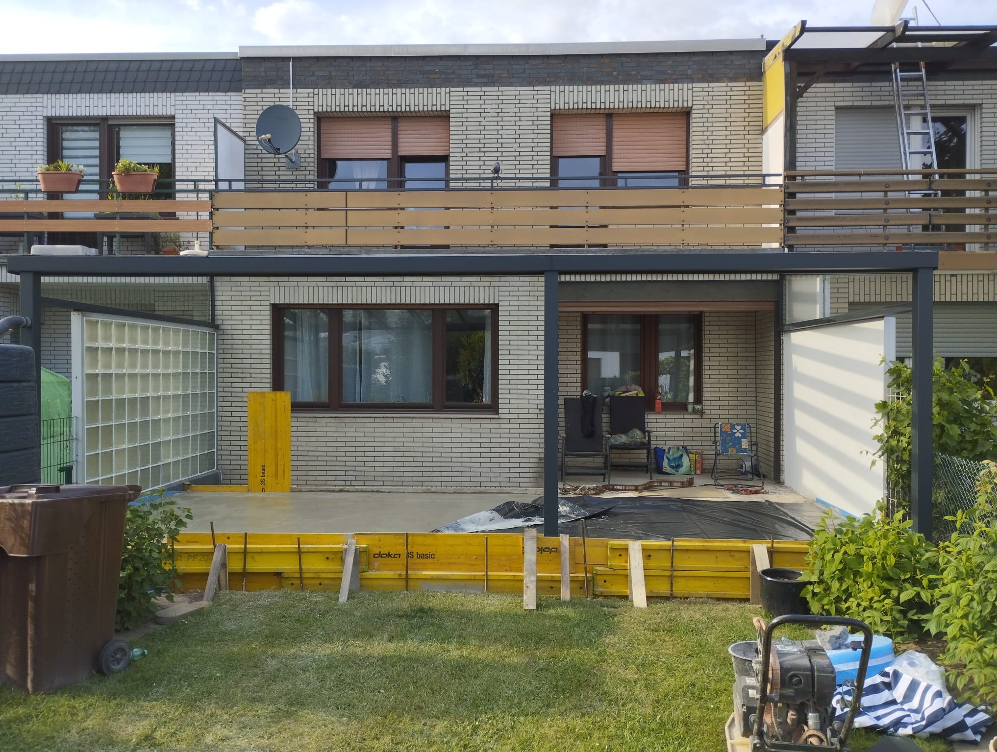 Budowa tarasu przy domu z cegły: żółte szalunki, w tle balkon z drewnianą balustradą, widoczny sprzęt budowlany i pojemnik na śmieci. Etap wylewania betonu.