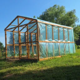 Drewniana szklarnia z folią na trawniku w słoneczny dzień. Wewnątrz widoczne sadzonki. Konstrukcja nowa, z elementami mocującymi folię. Tło: błękitne niebo i zieleń drzew.