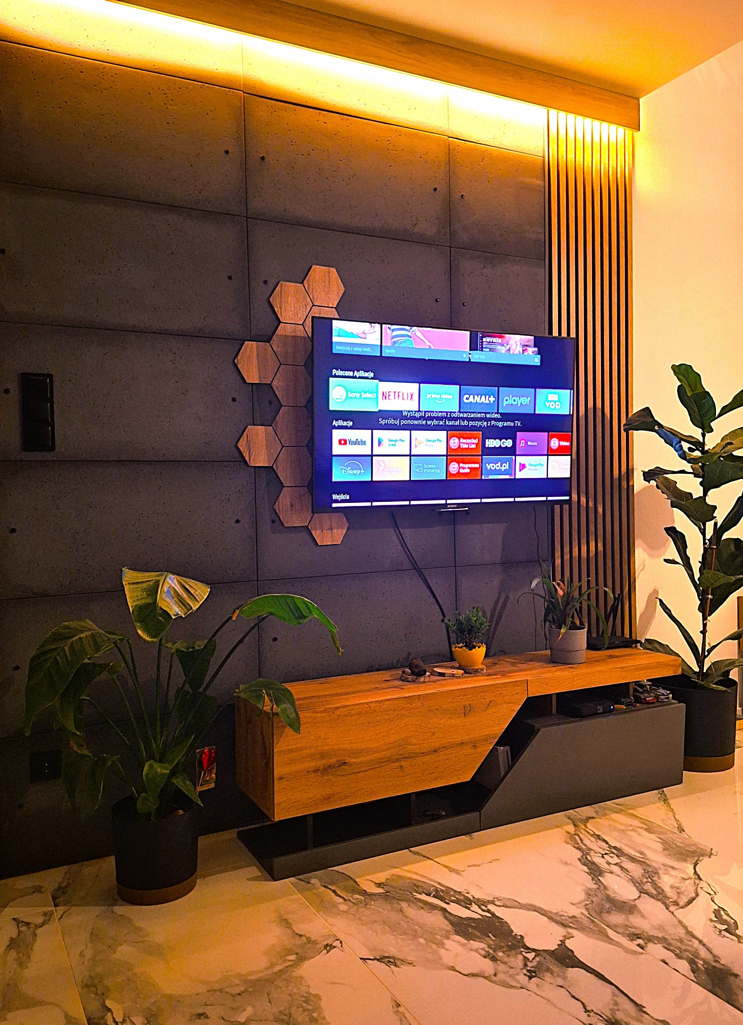 Nowoczesne wnętrze: ściana z betonowych płyt, drewniane heksagony, TV, lamelki, szafka RTV i rośliny doniczkowe, podświetlenie LED.