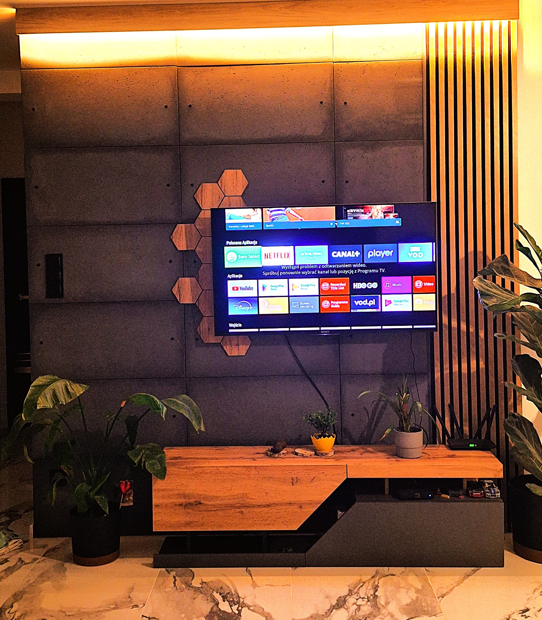 Nowoczesna ściana TV z betonowych paneli, drewnianych heksagonów i lameli. Podświetlenie LED. Szafka RTV z roślinami doniczkowymi. Marmurowa podłoga.