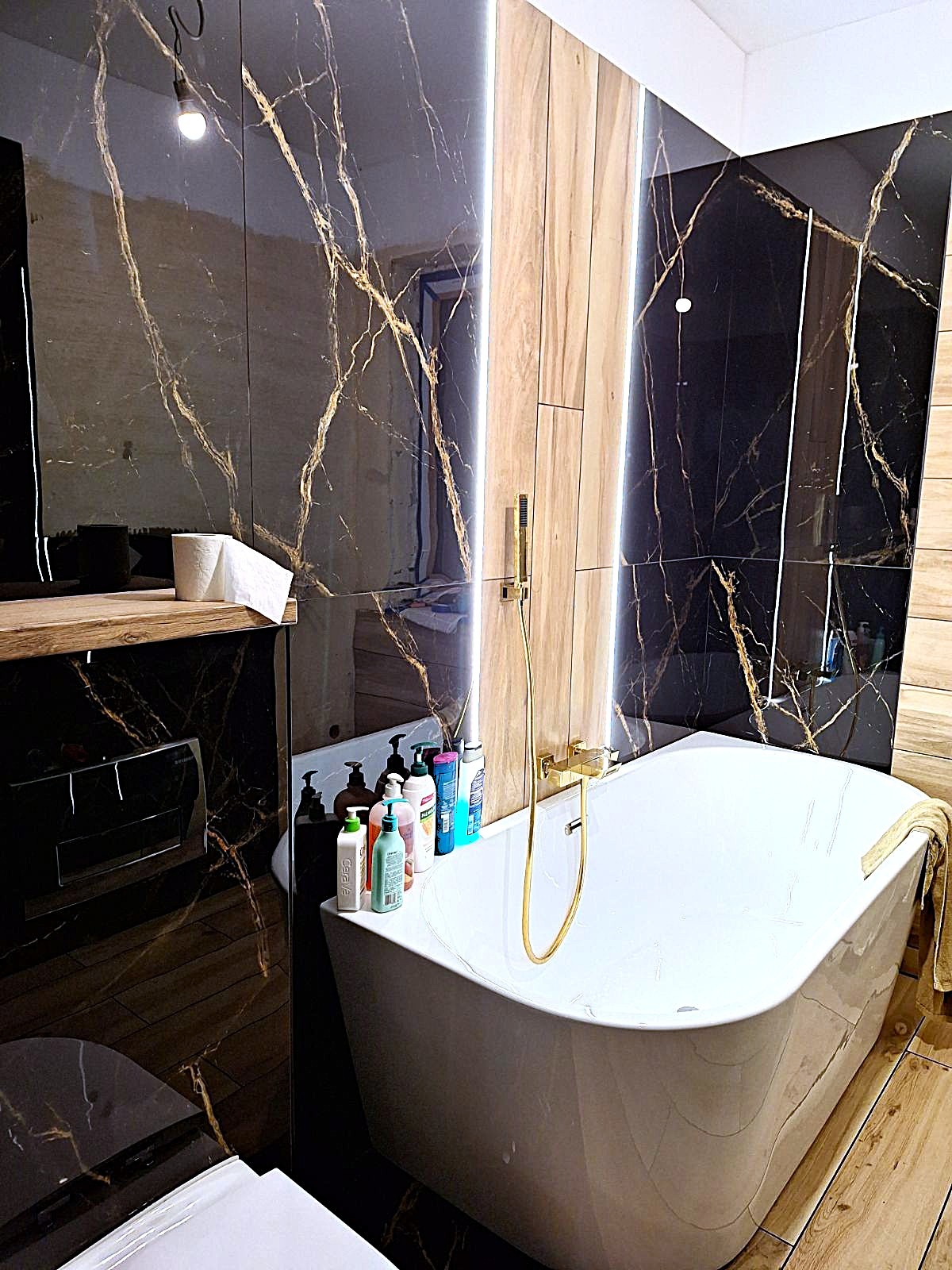 Nowoczesna łazienka z wanną wolnostojącą, czarnymi płytkami z żyłami złota i drewnianymi panelami z podświetleniem LED. Widoczne złote elementy armatury.