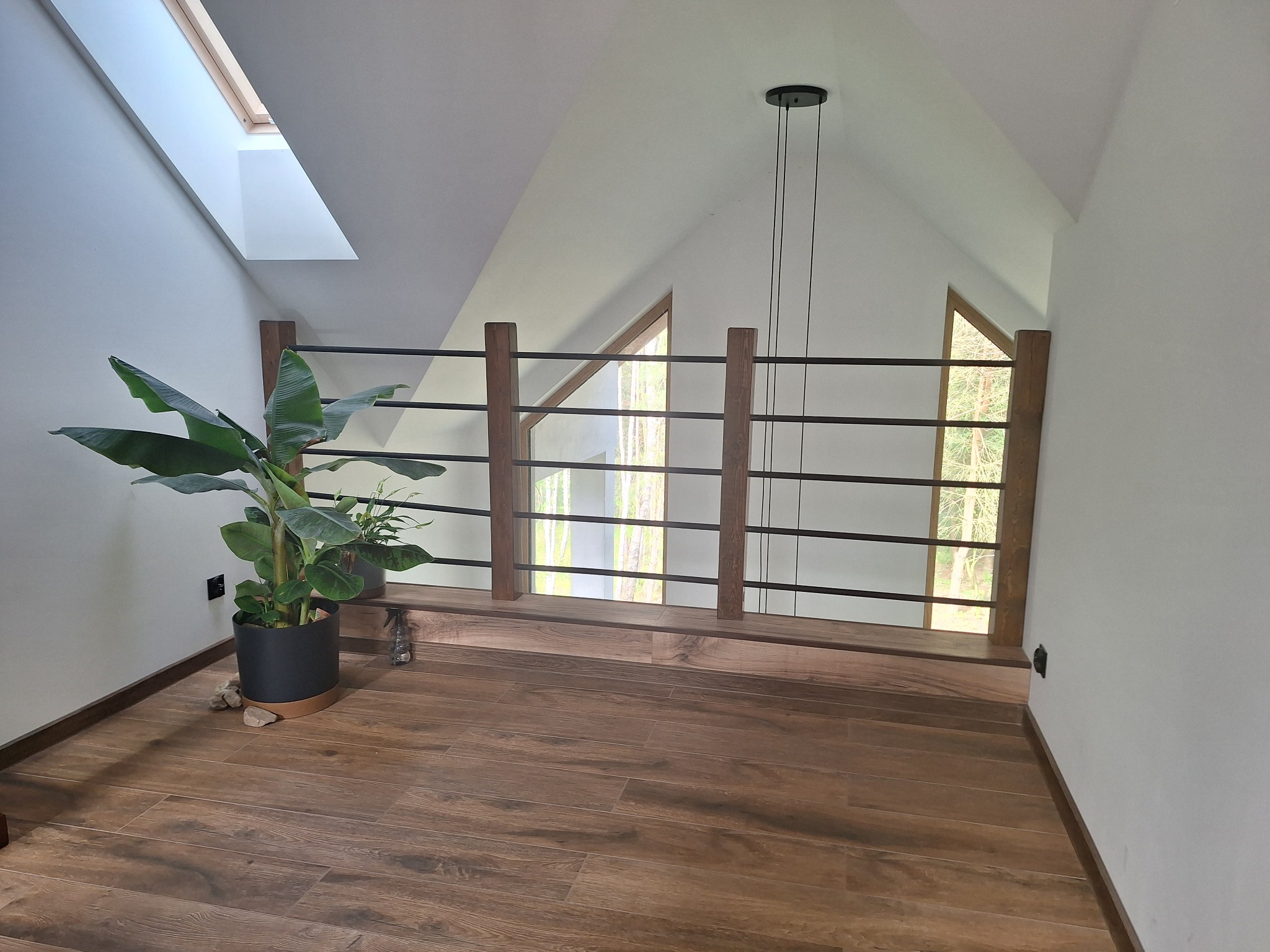 Podłoga z płytek drewnopodobnych na poddaszu, balustrada z drewnianymi słupkami i metalowymi prętami, okno dachowe, lampa wisząca i roślina doniczkowa.
