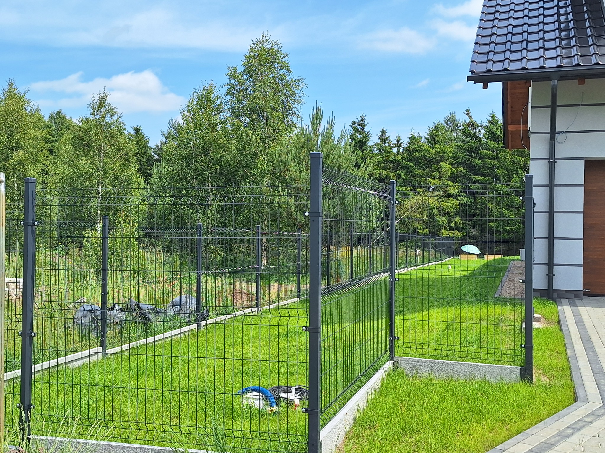 Nowoczesne ogrodzenie panelowe w kolorze grafitowym, otaczające zielony trawnik przy domu z ciemną dachówką. Widoczne słupki i siatka ogrodzeniowa.