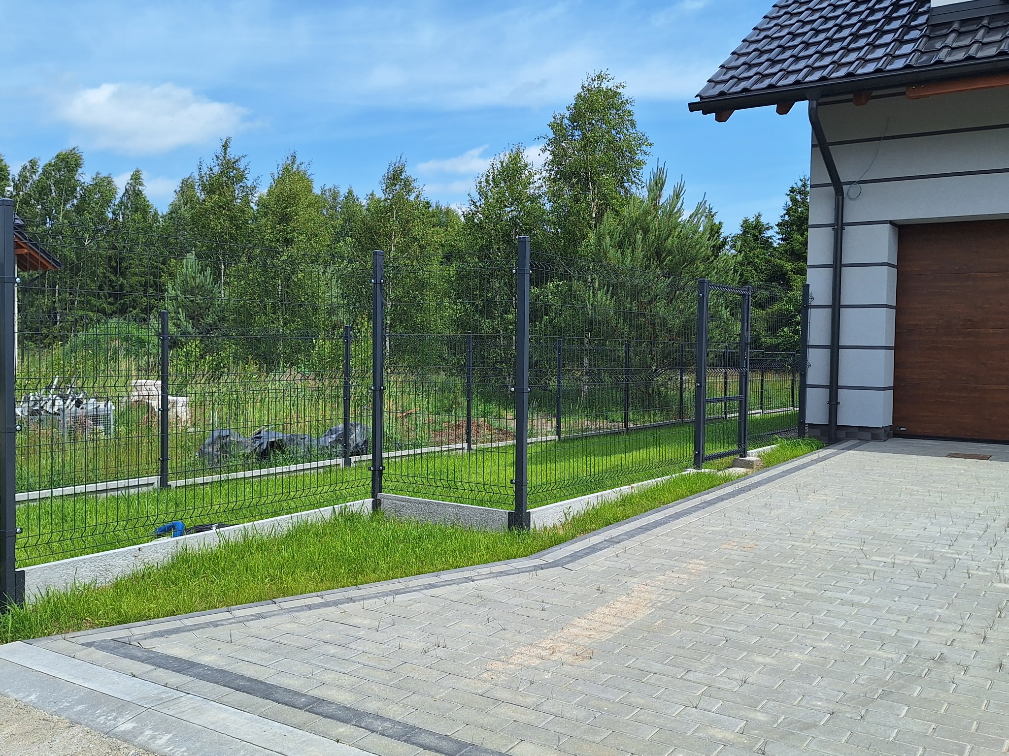 Nowoczesne ogrodzenie panelowe w kolorze grafitowym, oddzielające posesję od lasu, z widocznym podjazdem z kostki brukowej i fragmentem budynku z brązową bramą garażową.