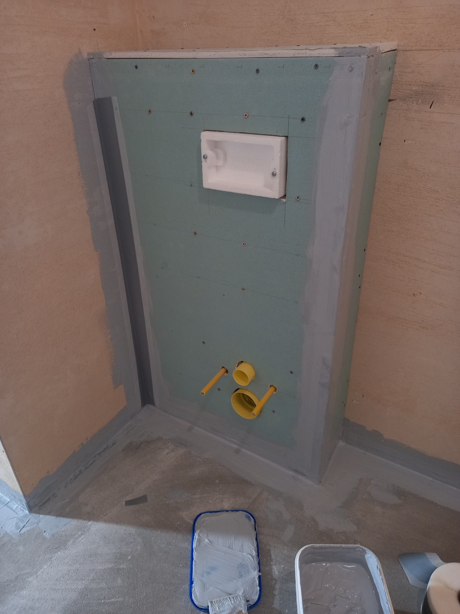 Przygotowanie ściany z płyt g-k pod zabudowę WC: widoczne profile, instalacja, zabezpieczenie hydroizolacją. Na podłodze narzędzia malarskie.