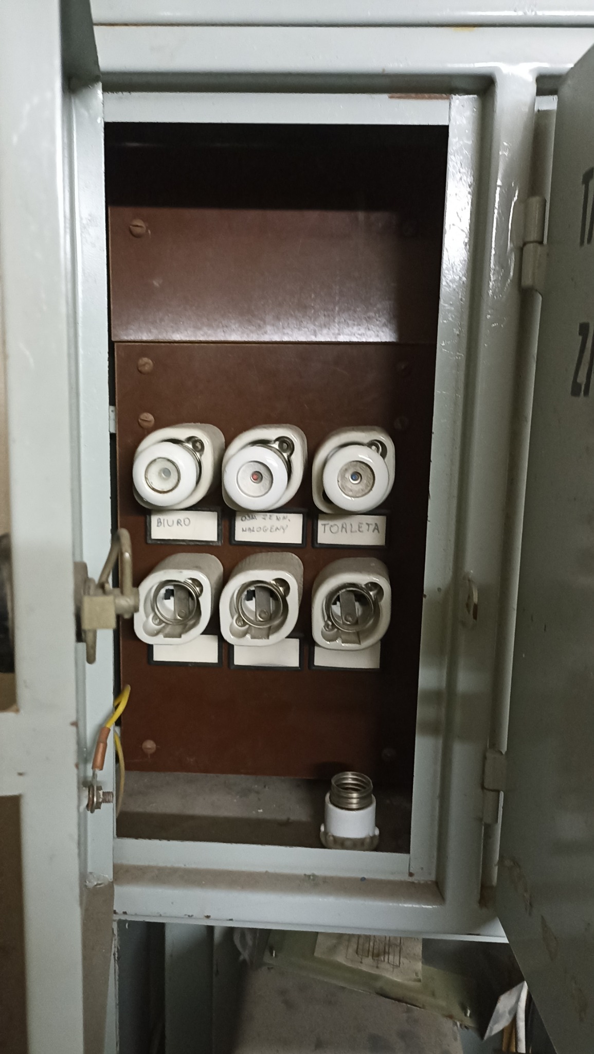 Otwarta, stara skrzynka bezpiecznikowa z ceramicznymi bezpiecznikami i odręcznymi etykietami: 'Biuro', 'Ośw. zewn. Halogeny', 'Toaleta'. Widoczny kabel uziemiający.