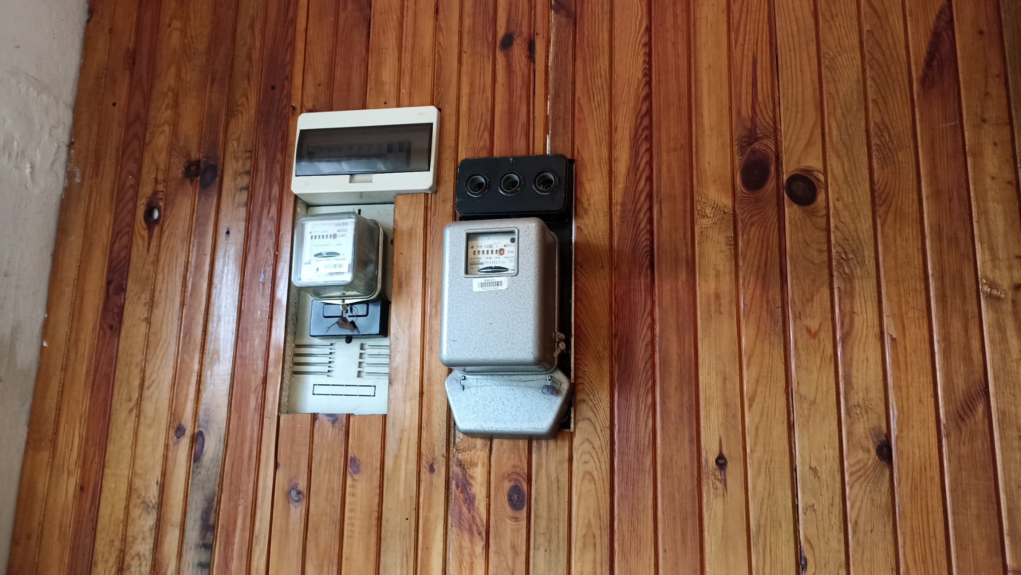 Dwa liczniki elektryczne, jeden biały, drugi szary, zamontowane na boazerii. Widoczna skrzynka bezpiecznikowa. Ujęcie z lekkiego dołu, oświetlenie dzienne.