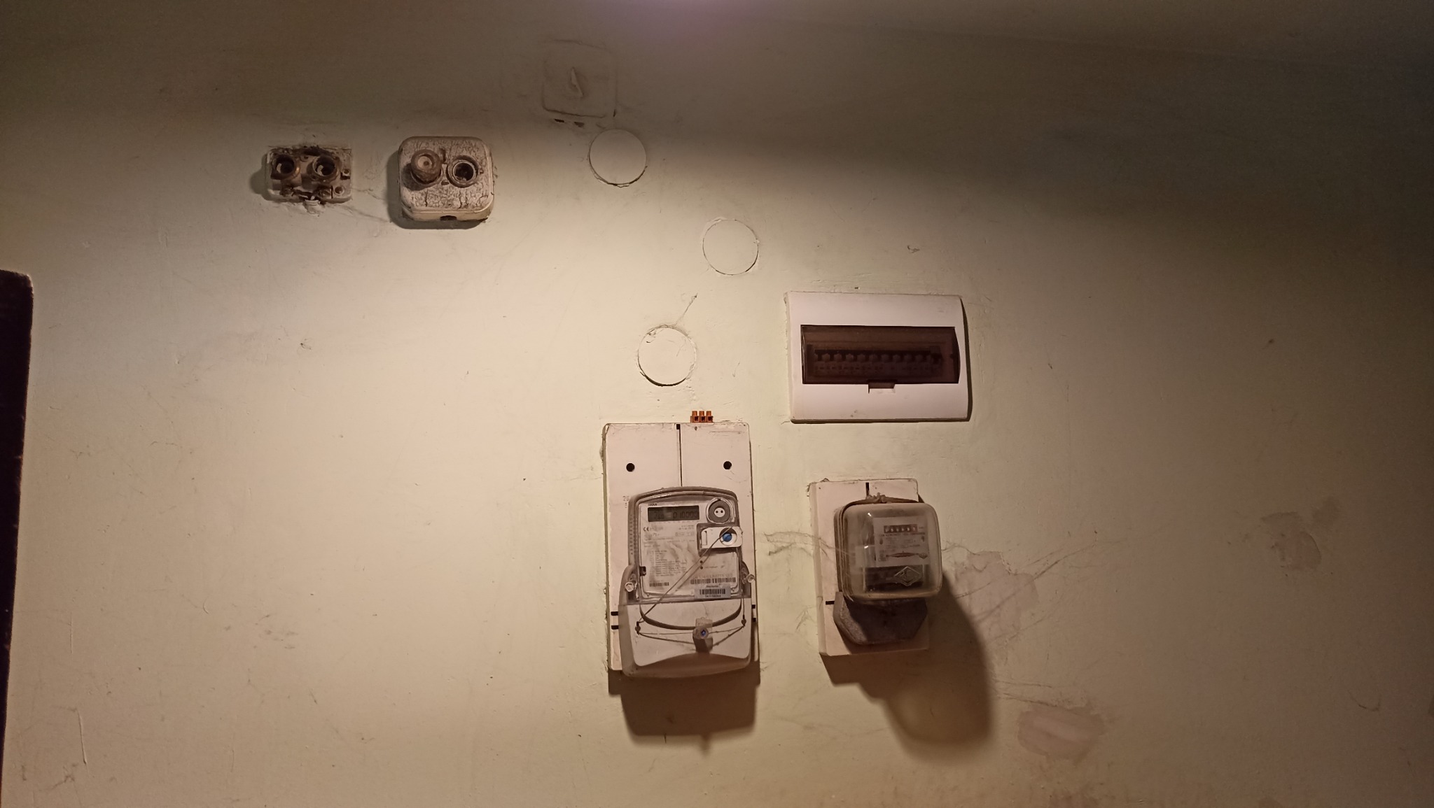 Stara instalacja elektryczna z dwoma licznikami, puszkami i skrzynką bezpiecznikową na ścianie w odcieniach beżu, z widocznymi śladami po wcześniejszych instalacjach.