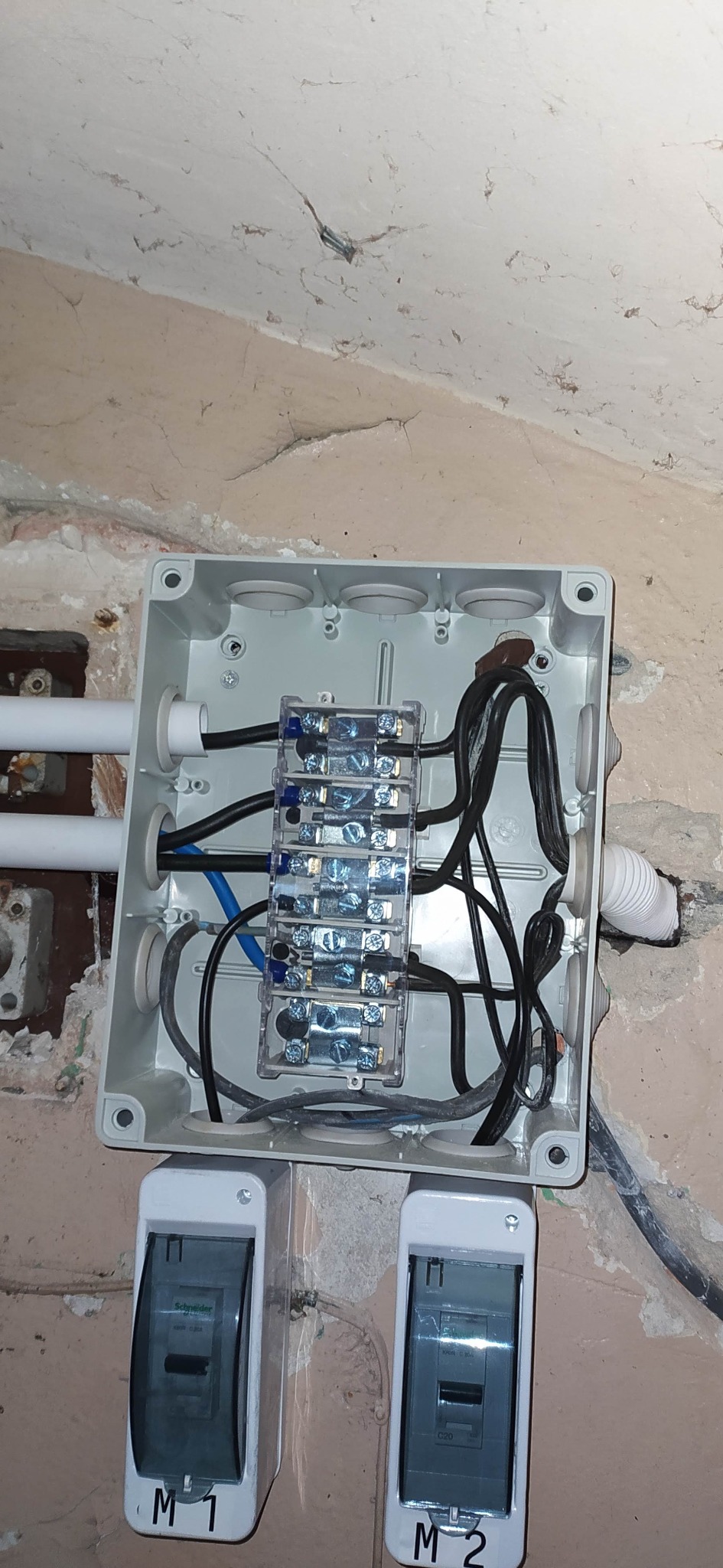 Otwarta skrzynka elektryczna z bezpiecznikami i przewodami na ścianie z widocznymi dwoma wyłącznikami nadprądowymi M1 i M2.