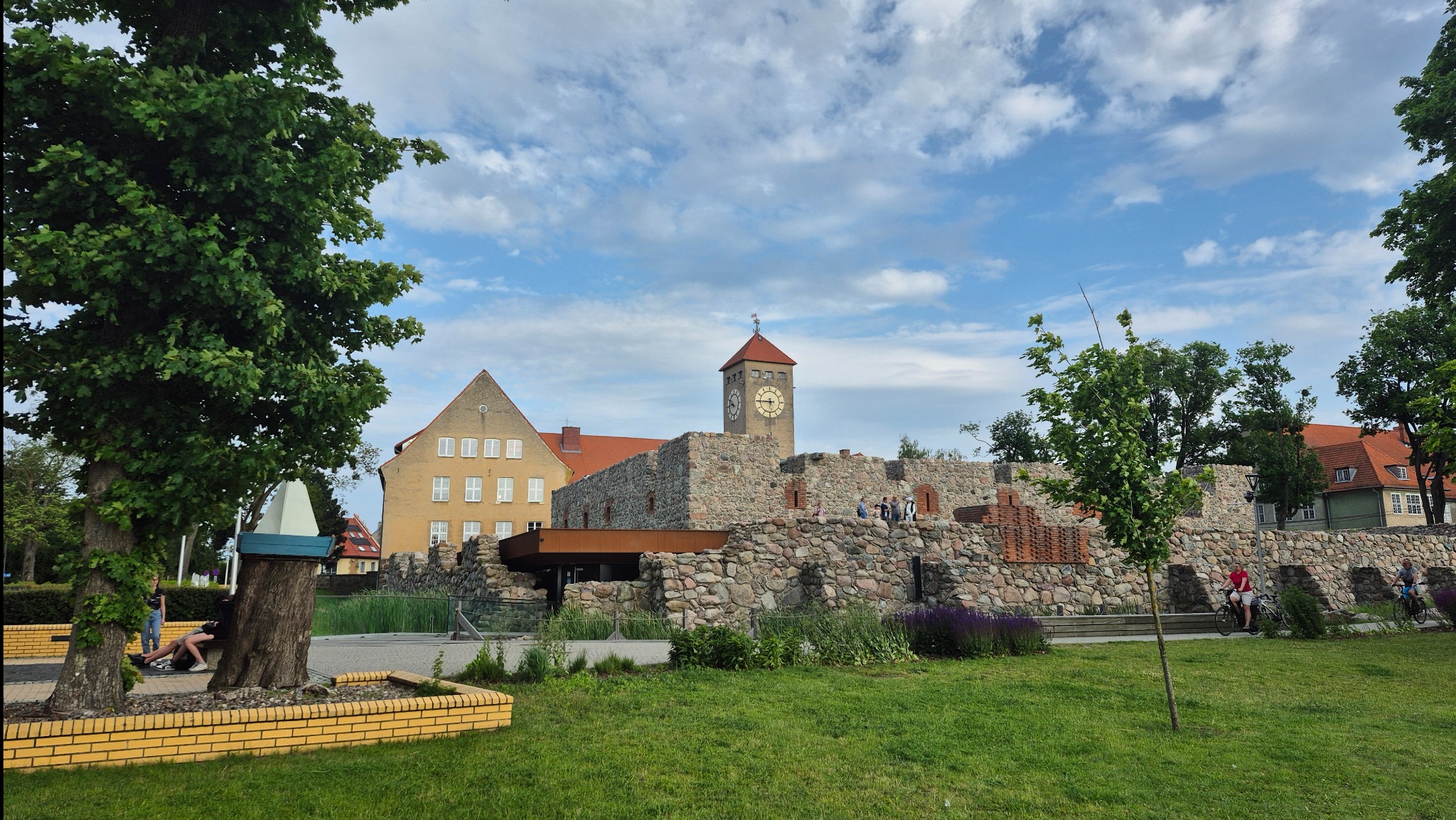 Szczytno: Zamek krzyżacki z zegarem i fragmentem parku w słoneczny dzień. Kamienna konstrukcja, ceglane detale, drzewa, osoby w tle. Ujęcie z poziomu trawnika.