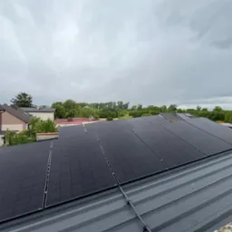 Panele fotowoltaiczne na szarym dachu blaszanej konstrukcji, widok z góry na tle zabudowań i pochmurnego nieba. Widoczne mocowania paneli.