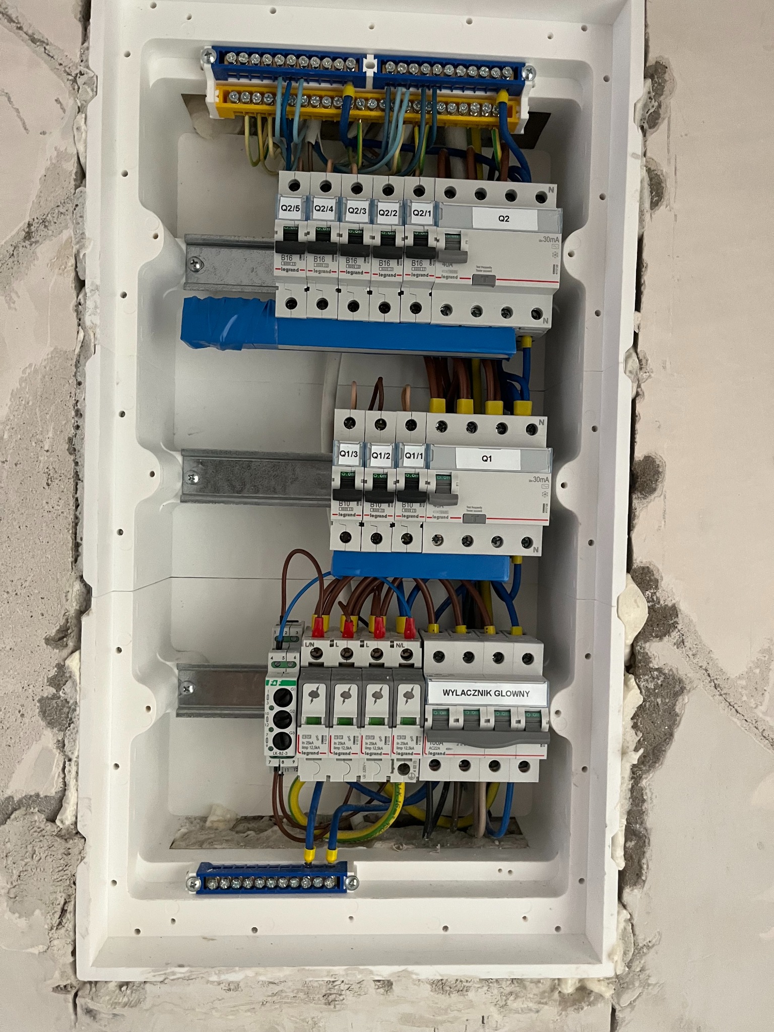Szczegółowe ujęcie skrzynki elektrycznej z zainstalowanymi bezpiecznikami i okablowaniem, w białej obudowie, w trakcie montażu w surowym murze.